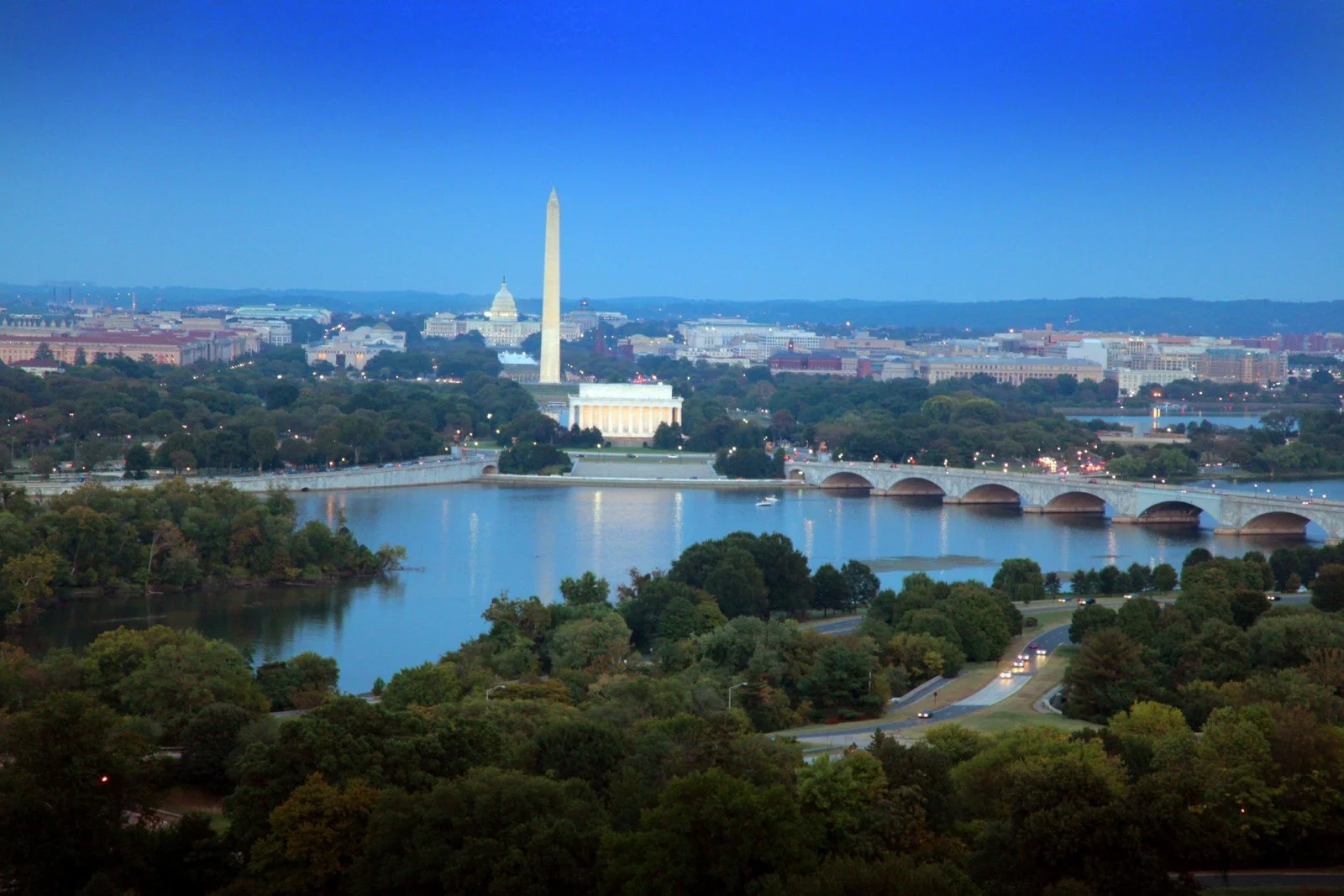 View of Washington D.C. skyline including the Washington Monument and Capitol building, with the Potomac River in the foreground and trees lining the riverbanks.