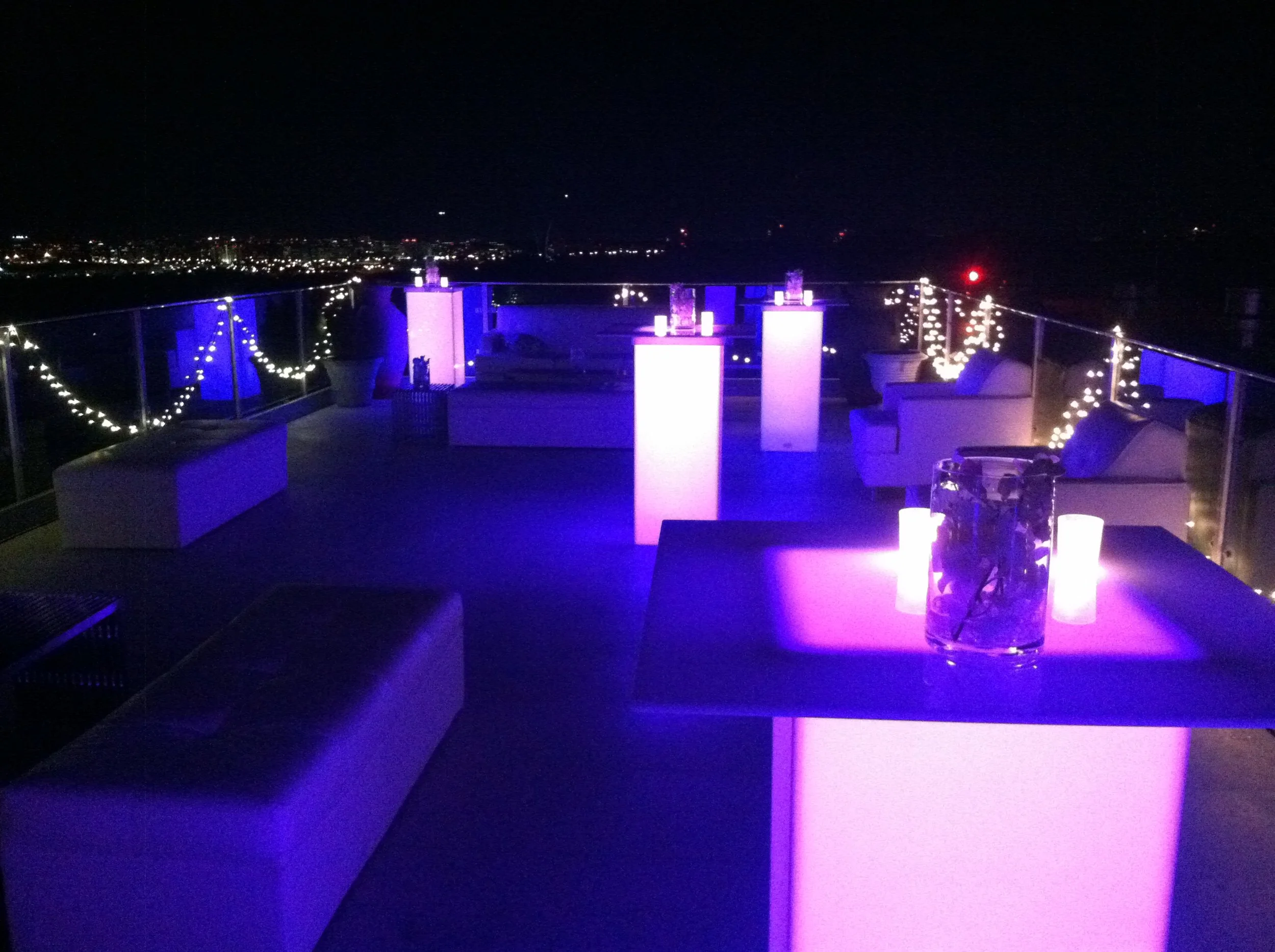 Rooftop party area at night decorated with purple and white lights, features lounge seating, tables with candles, cityscape view.