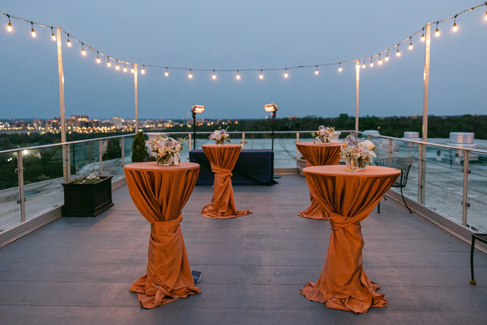 Outdoor rooftop reception area with four tall tables draped in orange tablecloths tied in a knot at the base, decorated with floral centerpieces, string lights overhead, and city lights in the distance.