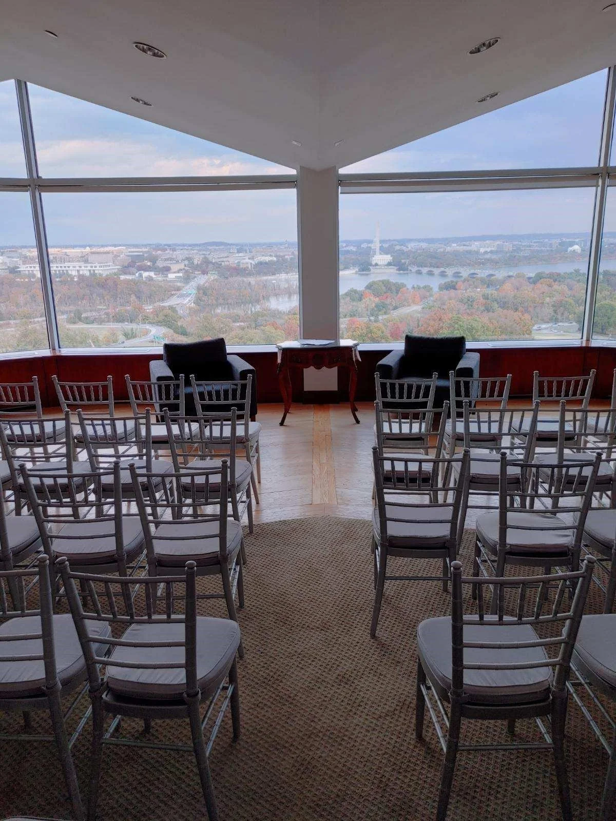 Room with chairs arranged for a ceremony, large windows showing a view of the city, river, and Washington Monument in the distance.