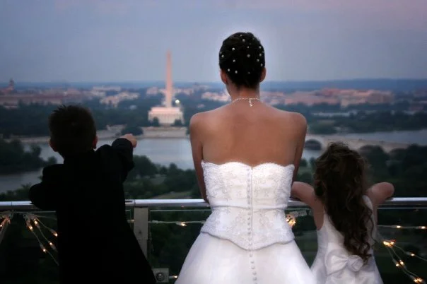 A woman with two children looking out over Washington D.C. from a high vantage point at sunset, with the Washington Monument visible in the background.