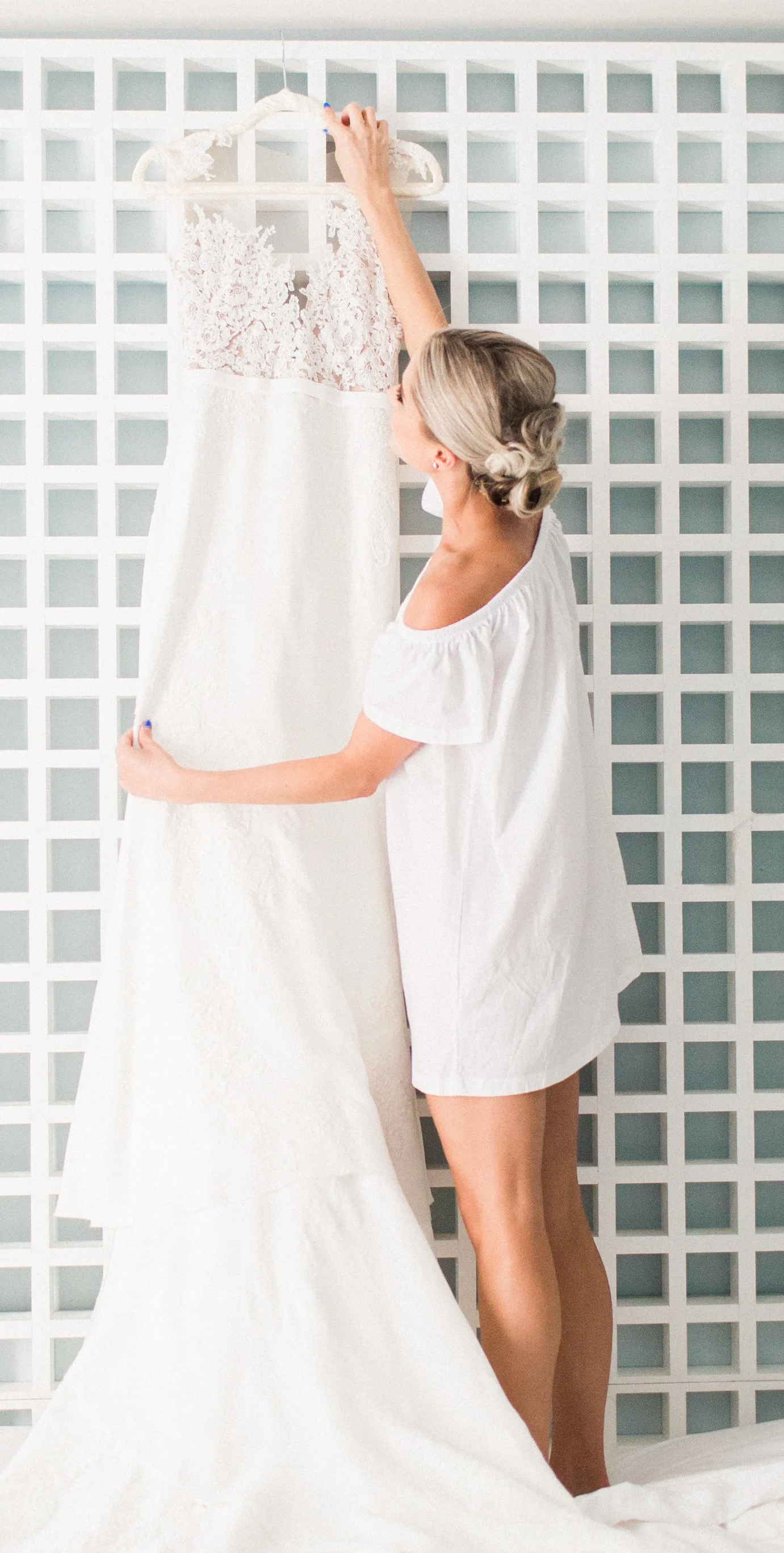 A woman with blonde hair styled in an updo is holding a white wedding dress with lace details on a hanger against a white grid wall.