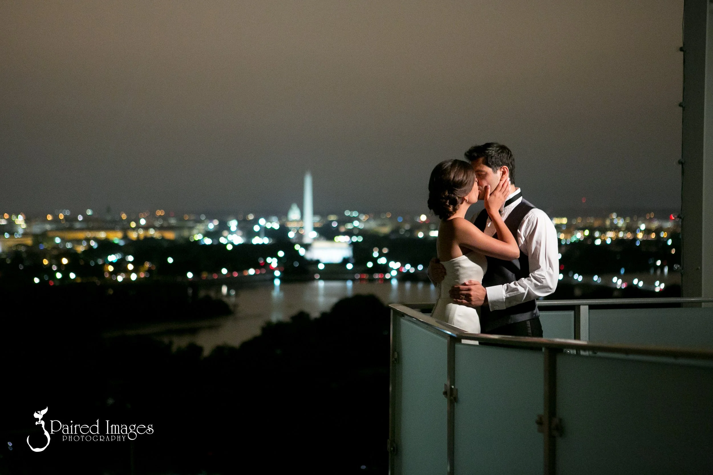 A couple sharing a kiss on a balcony at night, with blurred city lights and landmarks in the background.