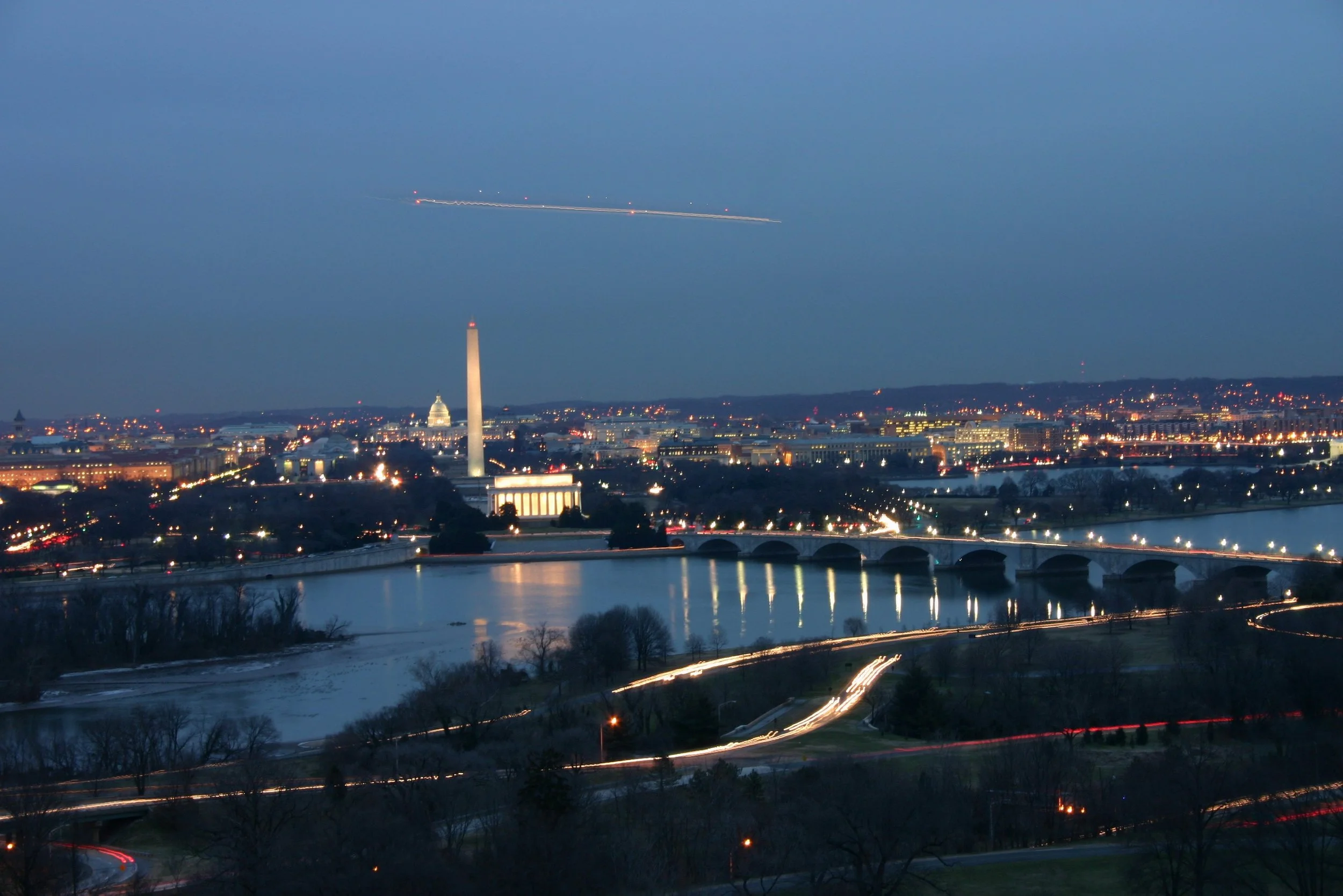 Nighttime cityscape of Washington D.C. with illuminated landmarks including the Lincoln Memorial, Washington Monument, and U.S. Capitol, along with a river and bridges, and aircraft trails in the blue sky.