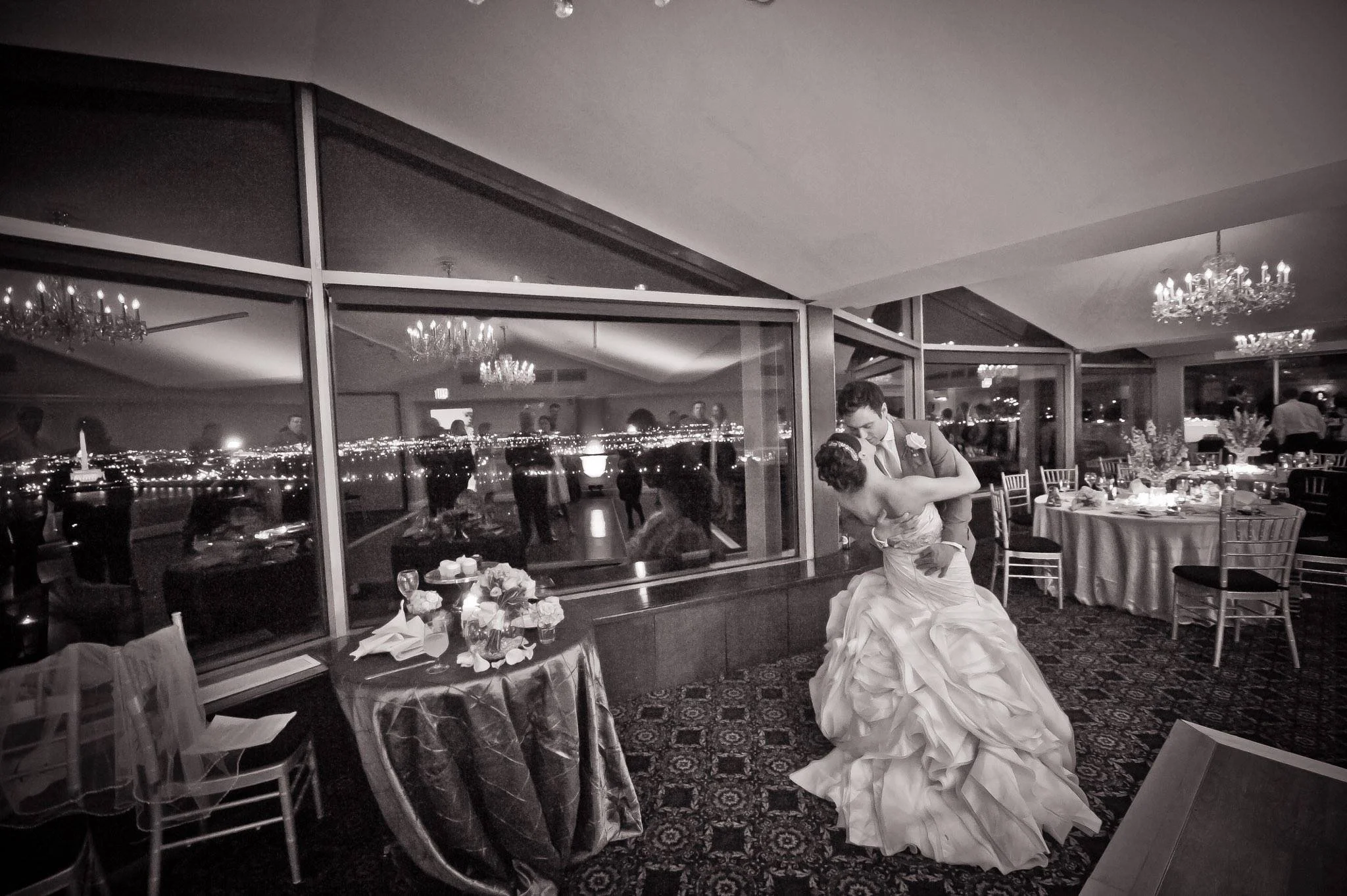 A couple dancing at a wedding reception in a decorated banquet hall at night, with city lights visible through large windows.