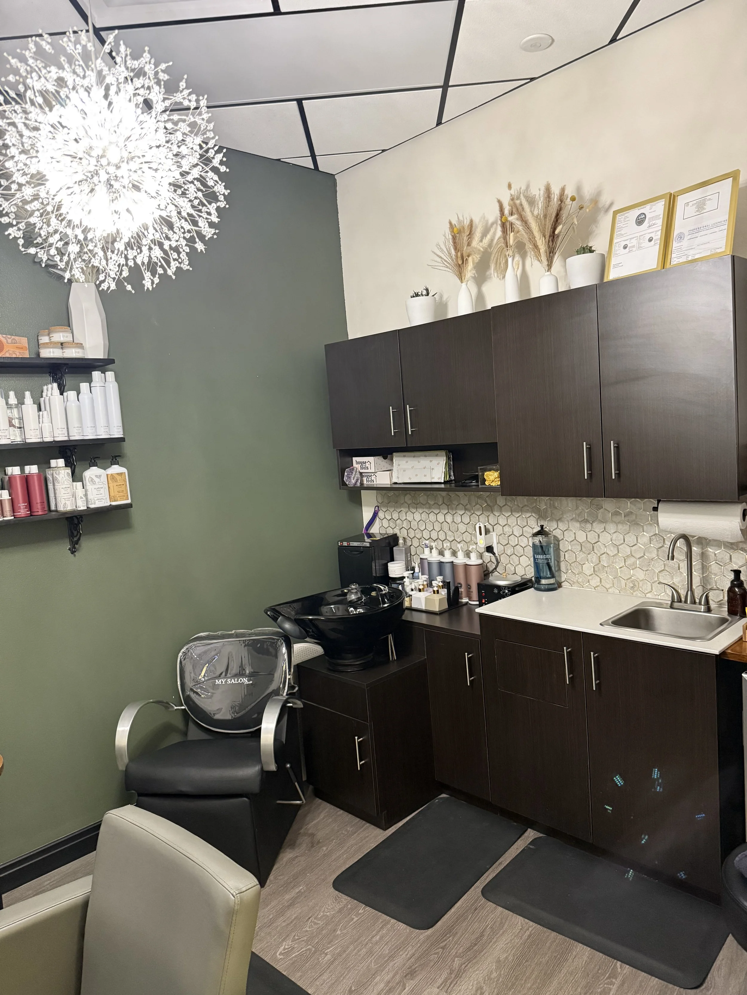 Interior view of a hair salon or beauty salon, featuring a black shampoo station with a chair, dark wooden cabinets, a sink, shelves with hair products, and decorative vases with dried plants. A bright chandelier hangs from the ceiling.