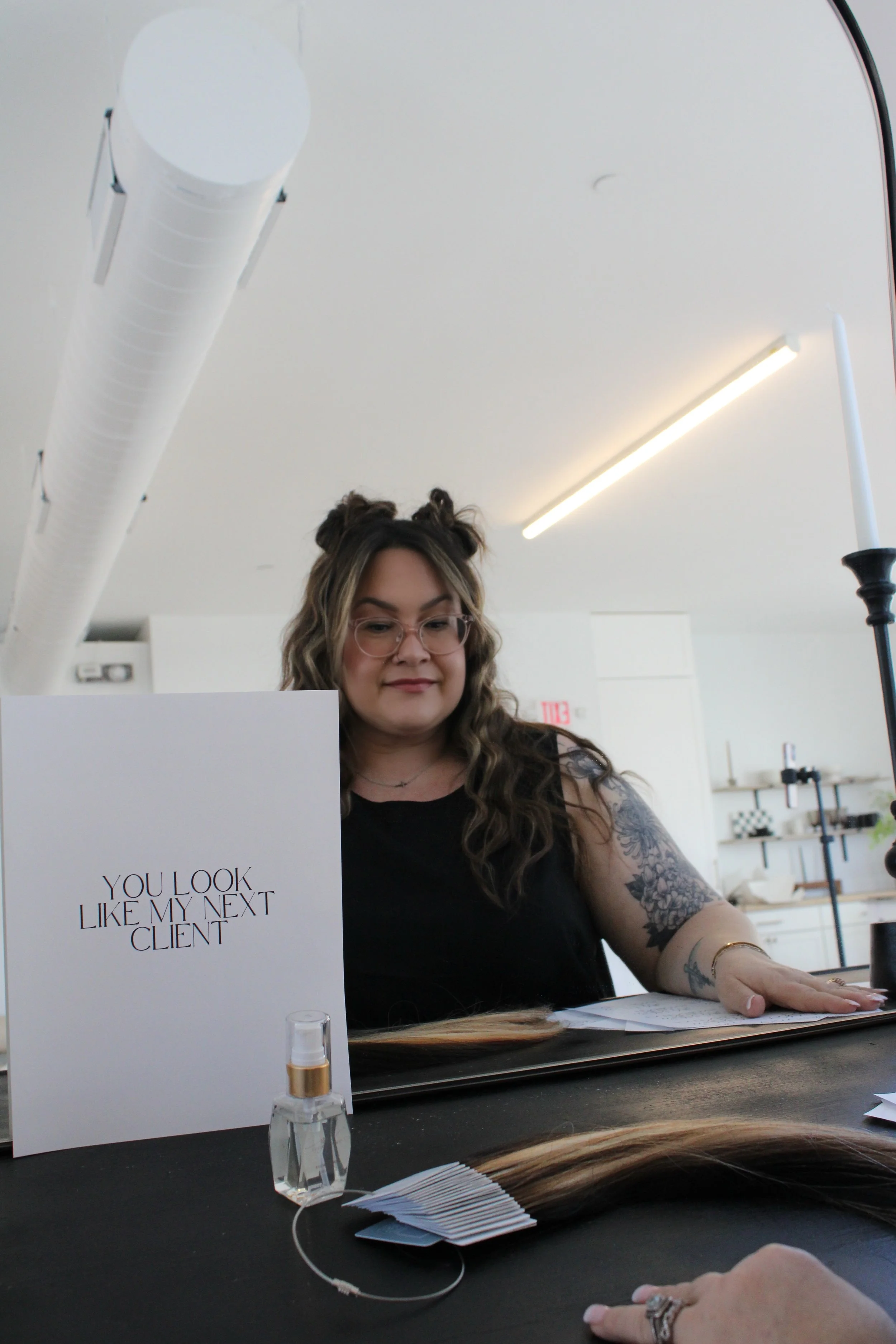 A woman with tattoos and glasses sitting at a table with hair samples, a sign that says "You look like my next client," and a spray bottle.