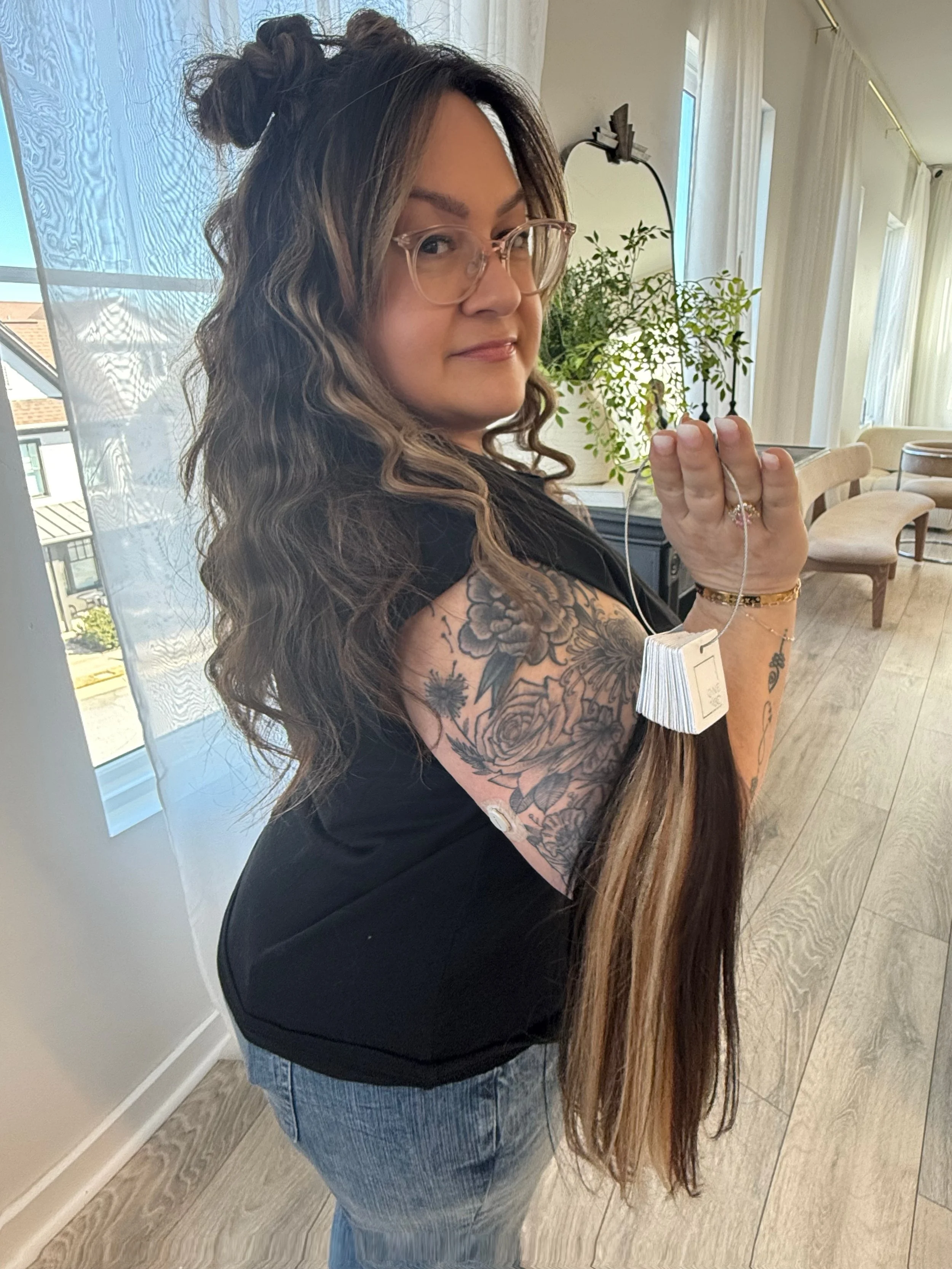 A woman with long, wavy hair and glasses is holding a stack of tea bags in her hand while standing indoors near a window with white curtains, a plant, and modern furniture.
