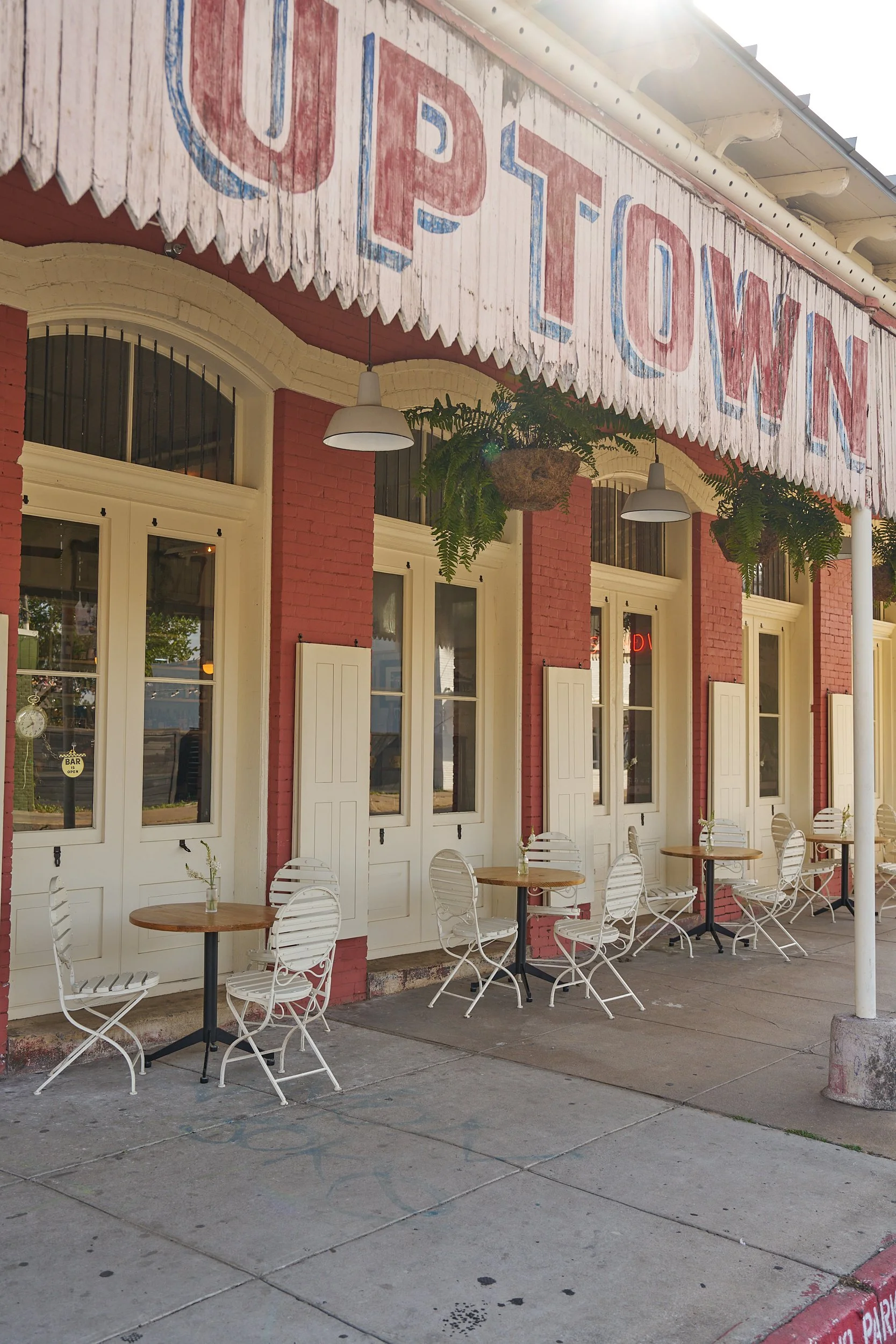Exterior of a vintage-style café with white tables and chairs outside, large windows, red brick walls, white shutters, hanging potted plants, and a weathered sign that reads 'UP TOWN'.