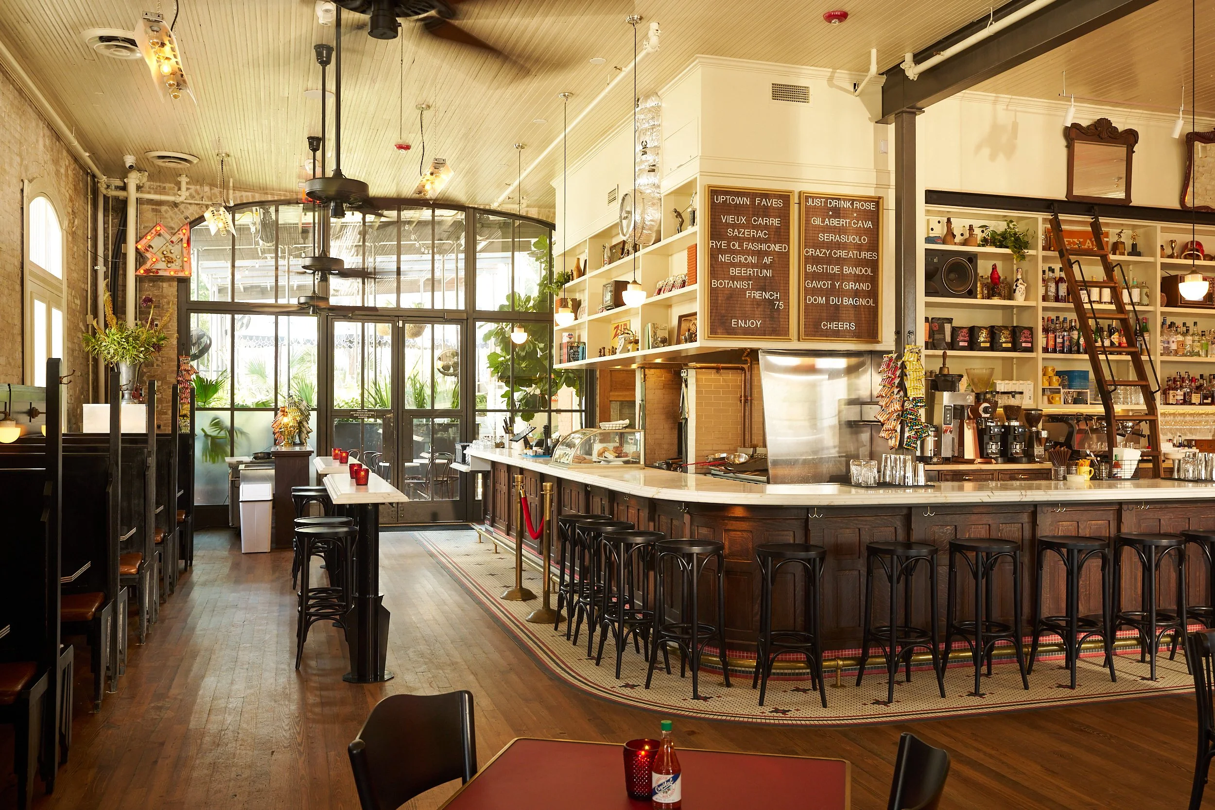 Interior of a cozy cafe with a curved wooden counter, black barstools, and a chalkboard menu. Natural light streams through large windows, and vintage-style decorations and plants adorn the space.