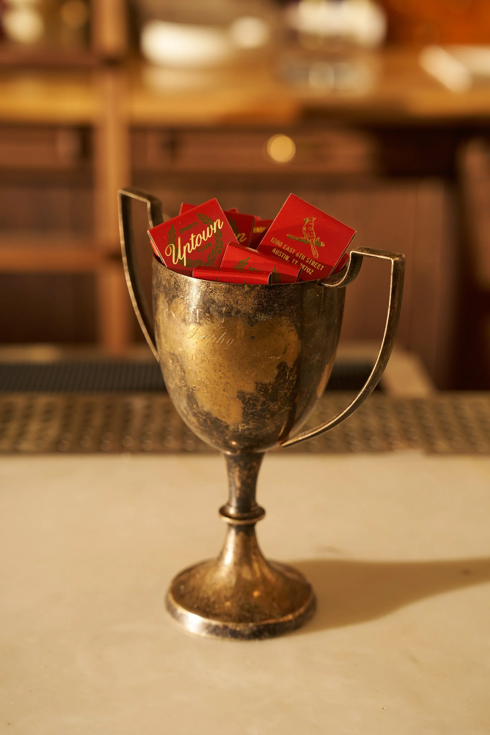 A vintage metal trophy cup filled with red matchbooks for Uptown in austin, placed on a light-colored surface in a warmly lit indoor setting.