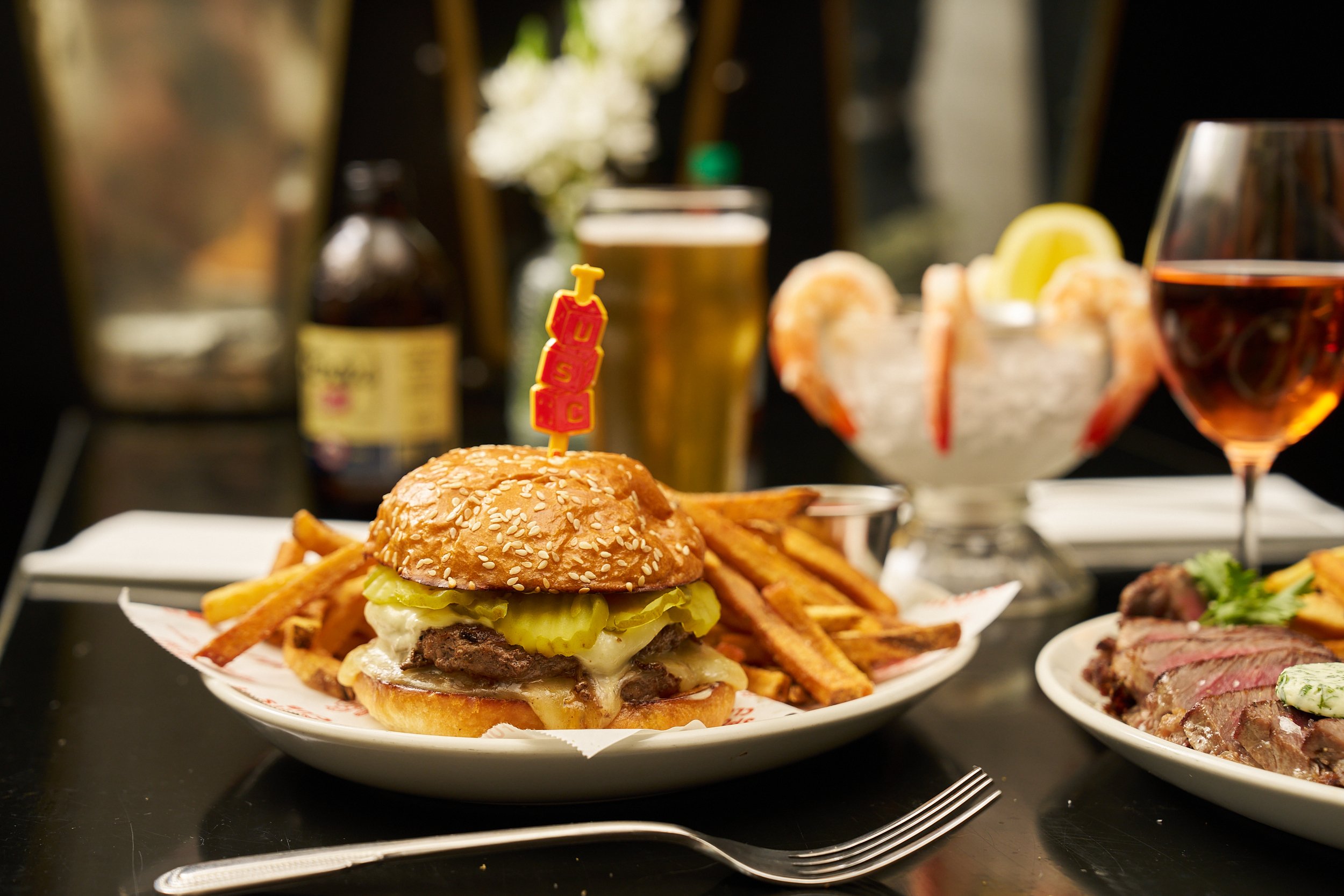 A cheeseburger with pickles, lettuce, and cheese topped with a 'USC' flag pick on a plate with French fries, accompanied by a glass of rosé wine, a bowl of shrimp with lemon slices, and a plate of sliced steak with herbs, on a black table at a restaurant.