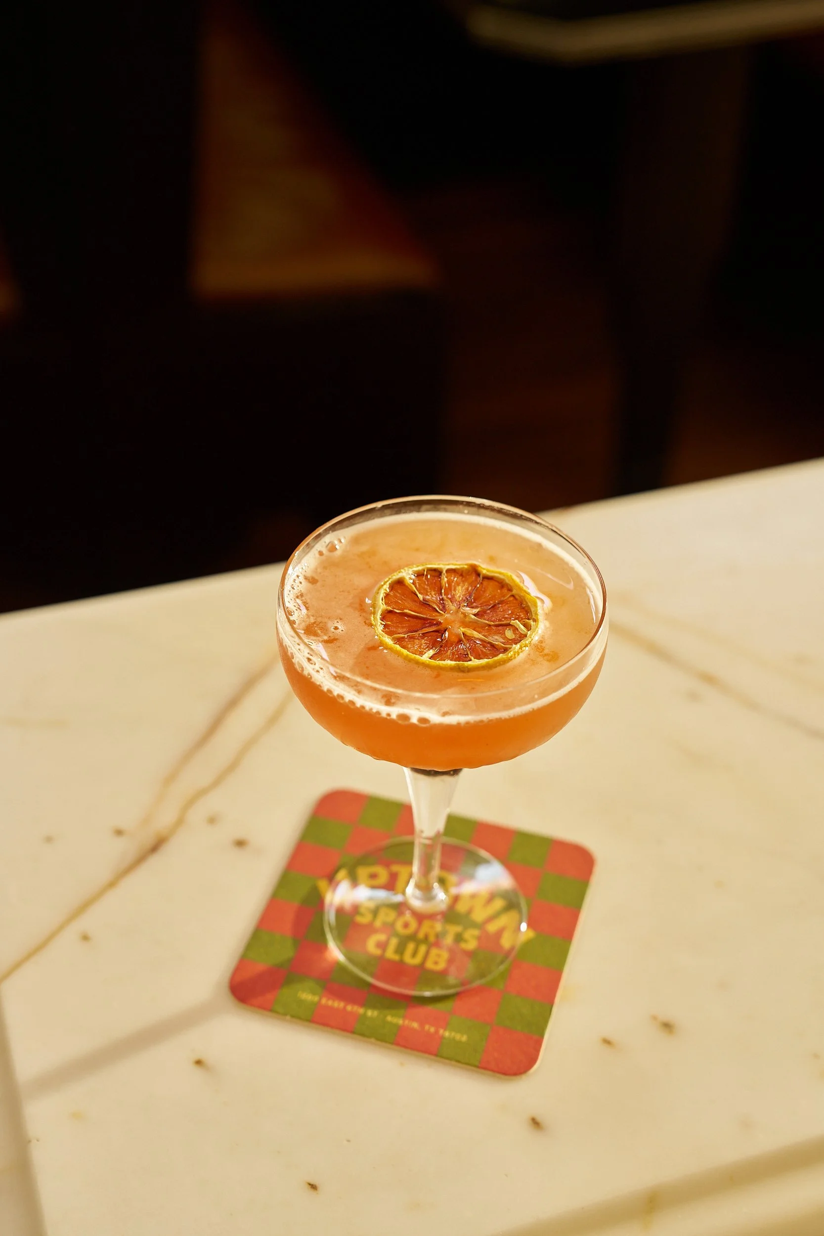 A pink cocktail garnished with a dried citrus slice, placed on a checkered coaster on a white marble table.