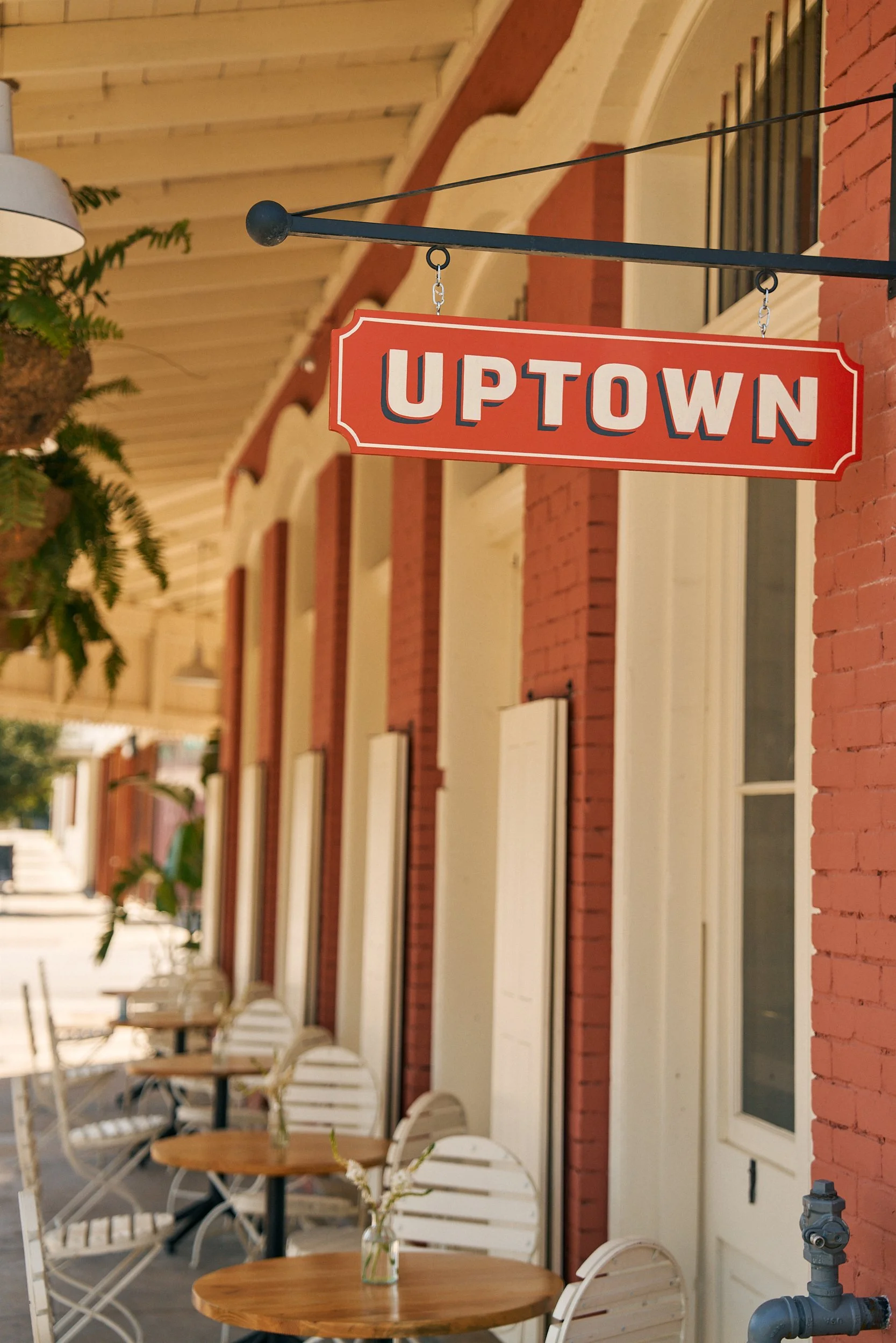 Street-side patio with round wooden tables, white chairs, potted plants, and a red sign hanging from the building that reads 'Uptown'.