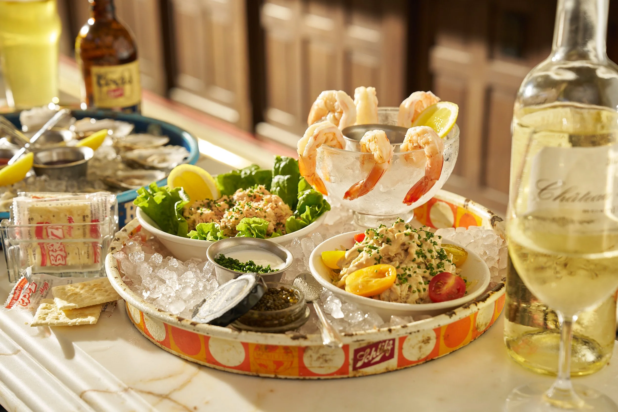 Seafood platter with shrimp, oyster crackers, lemon wedges, potato salad, and various condiments served on a bed of ice with a glass of white wine.