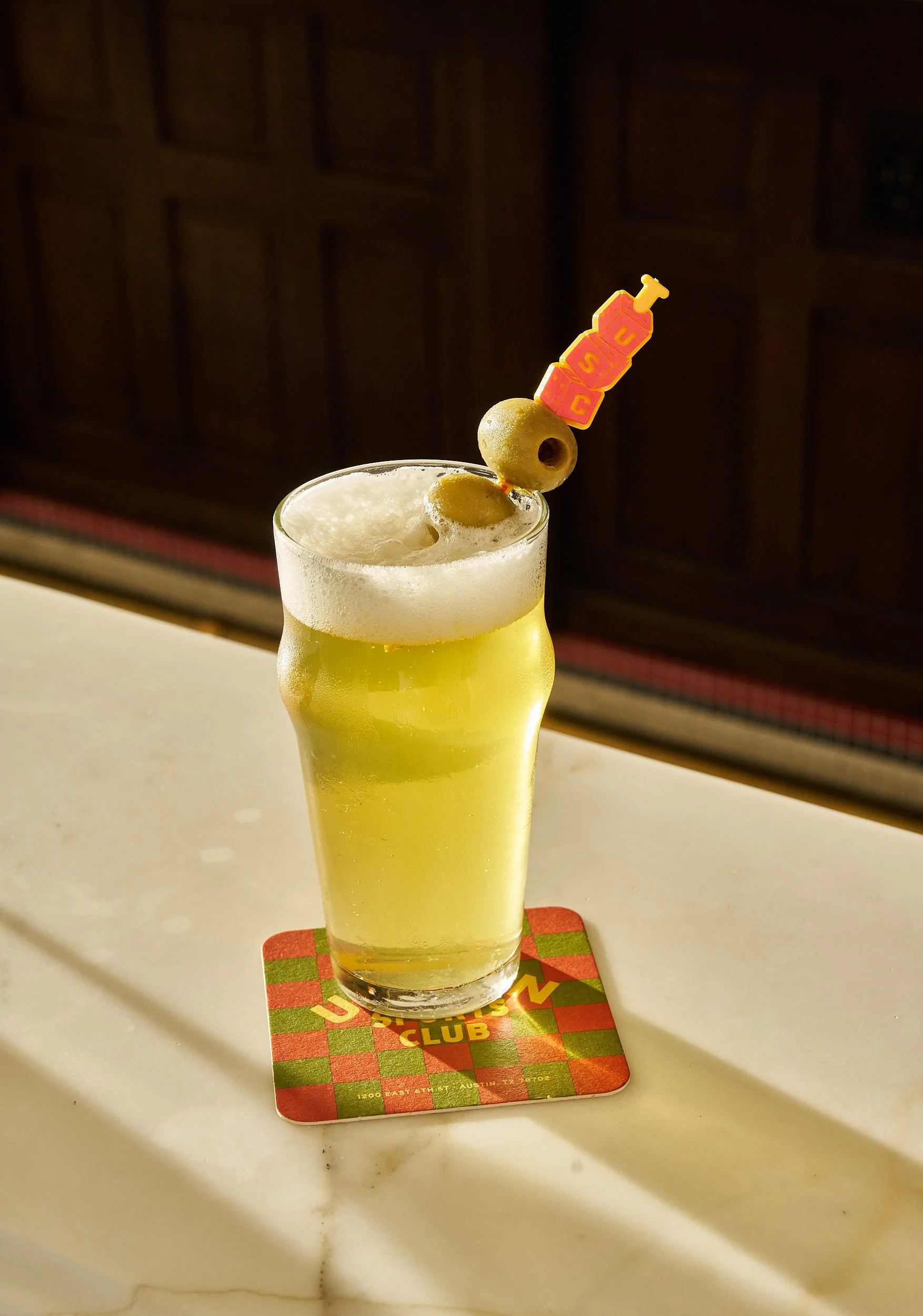 A glass of yellowish drink garnished with green olives and a pick with a pink and orange block design with the word "USC" on top of a red and green checkered coaster.