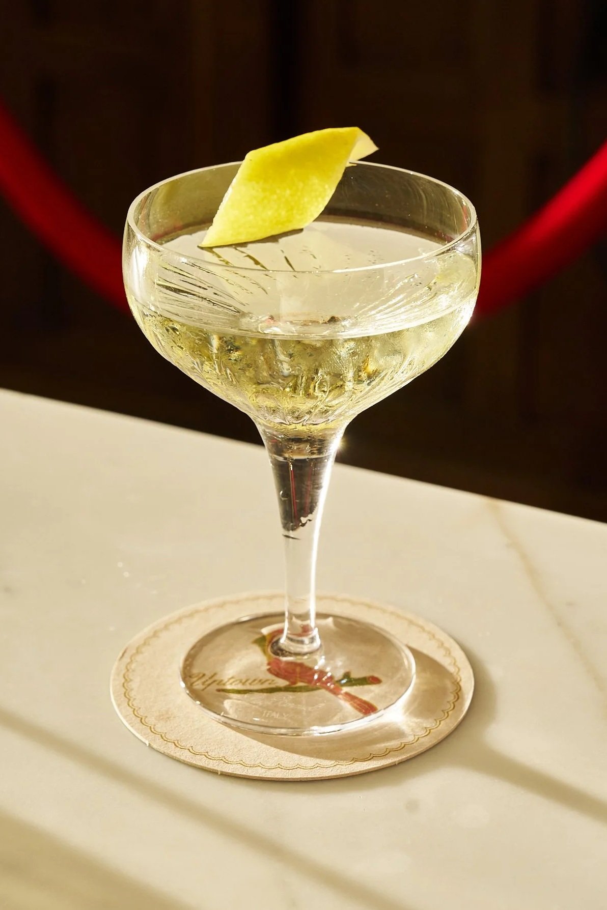 A cocktail glass with a slice of lemon garnish and a clear drink inside, placed on a coaster on a white surface.