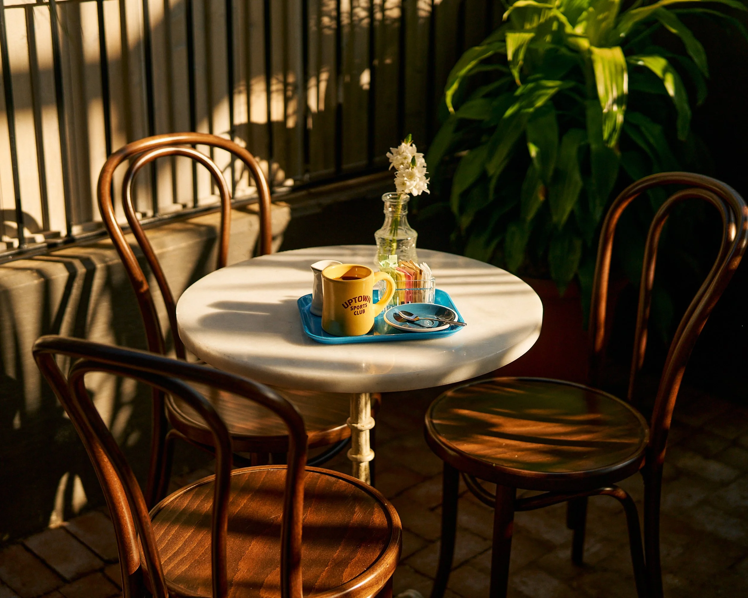 A small round white table set outdoors with three wooden chairs around it. The table has a blue tray with a yellow mug, a glass bottle with white flowers, a small plate, and sugar packets. Sunlight casts shadows on the table and chairs, with a large green plant in the background.