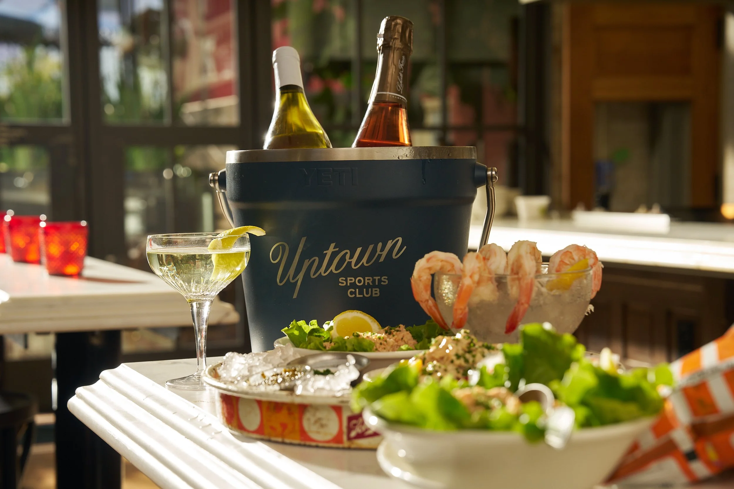 Bar table with seafood, salad, and drinks, including a bucket of bottled wine or beer, a glass of white wine with lemon, and a bowl of shrimp, set in a bright indoor space with a window.