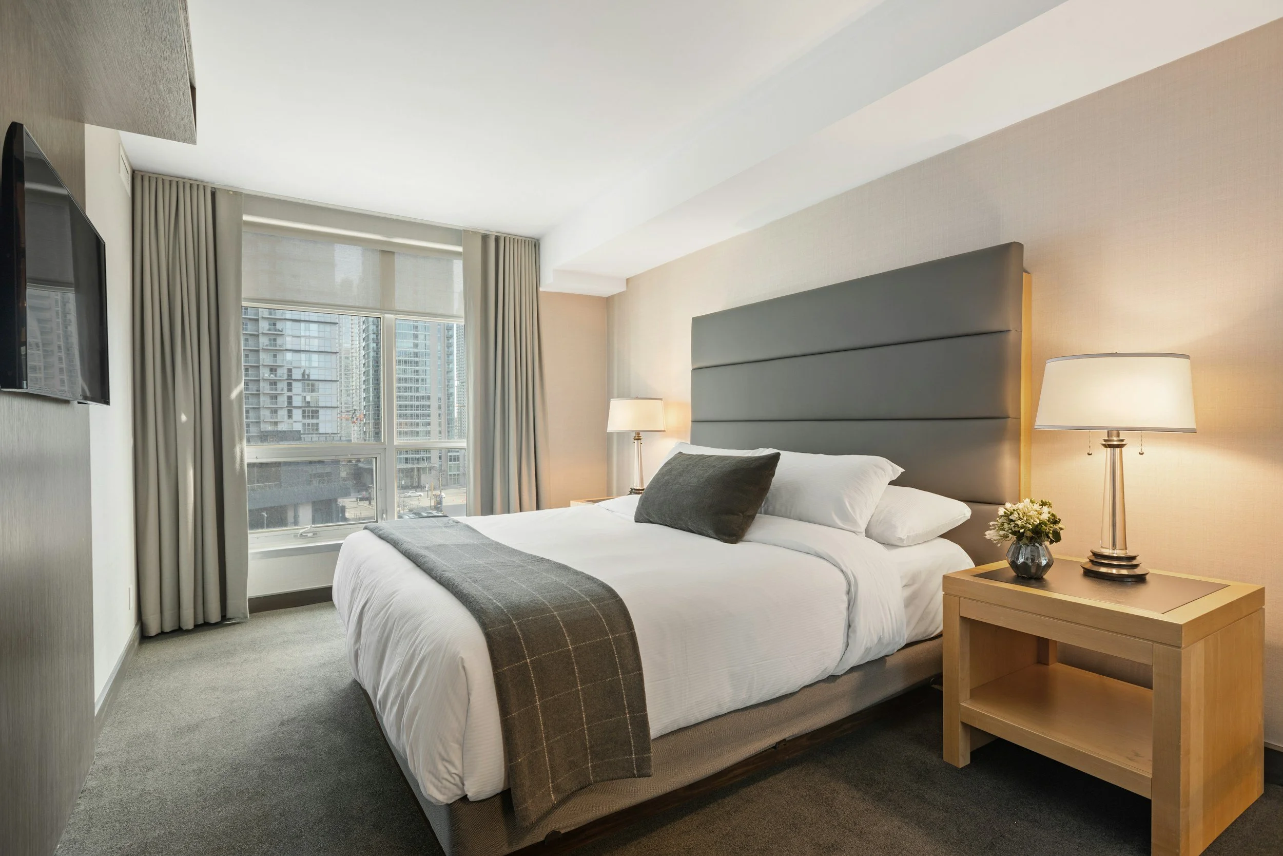 Hotel room with a large bed, gray headboard, two nightstands with lamps, flower vase, and a window showing city buildings.