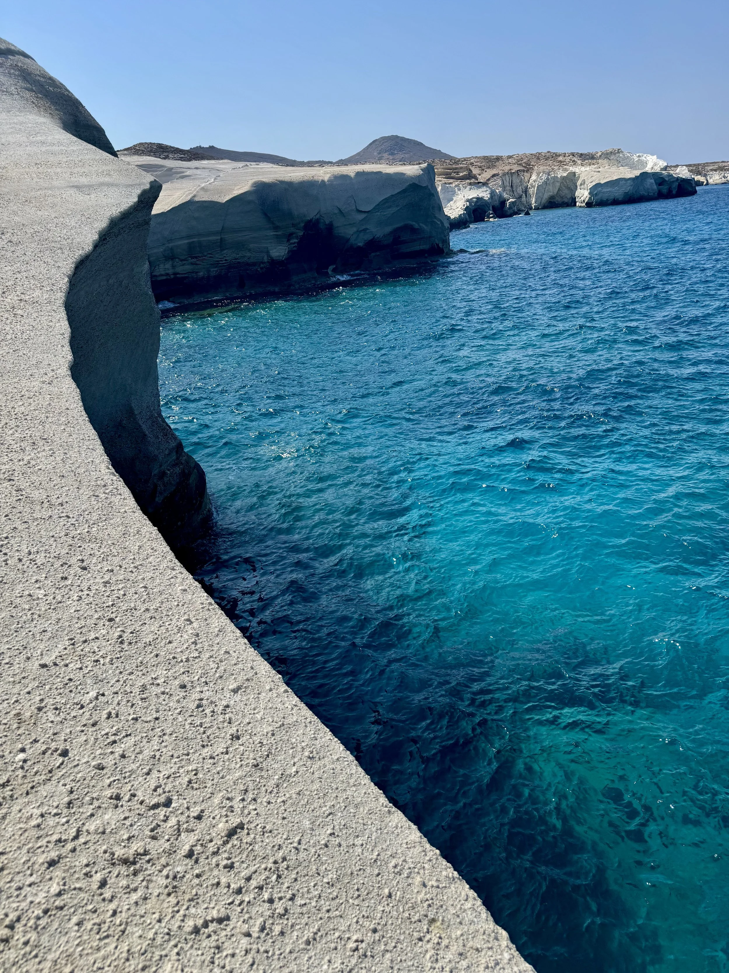 Cliffs with white and dark rock formations overlooking bright blue ocean water on a sunny day.