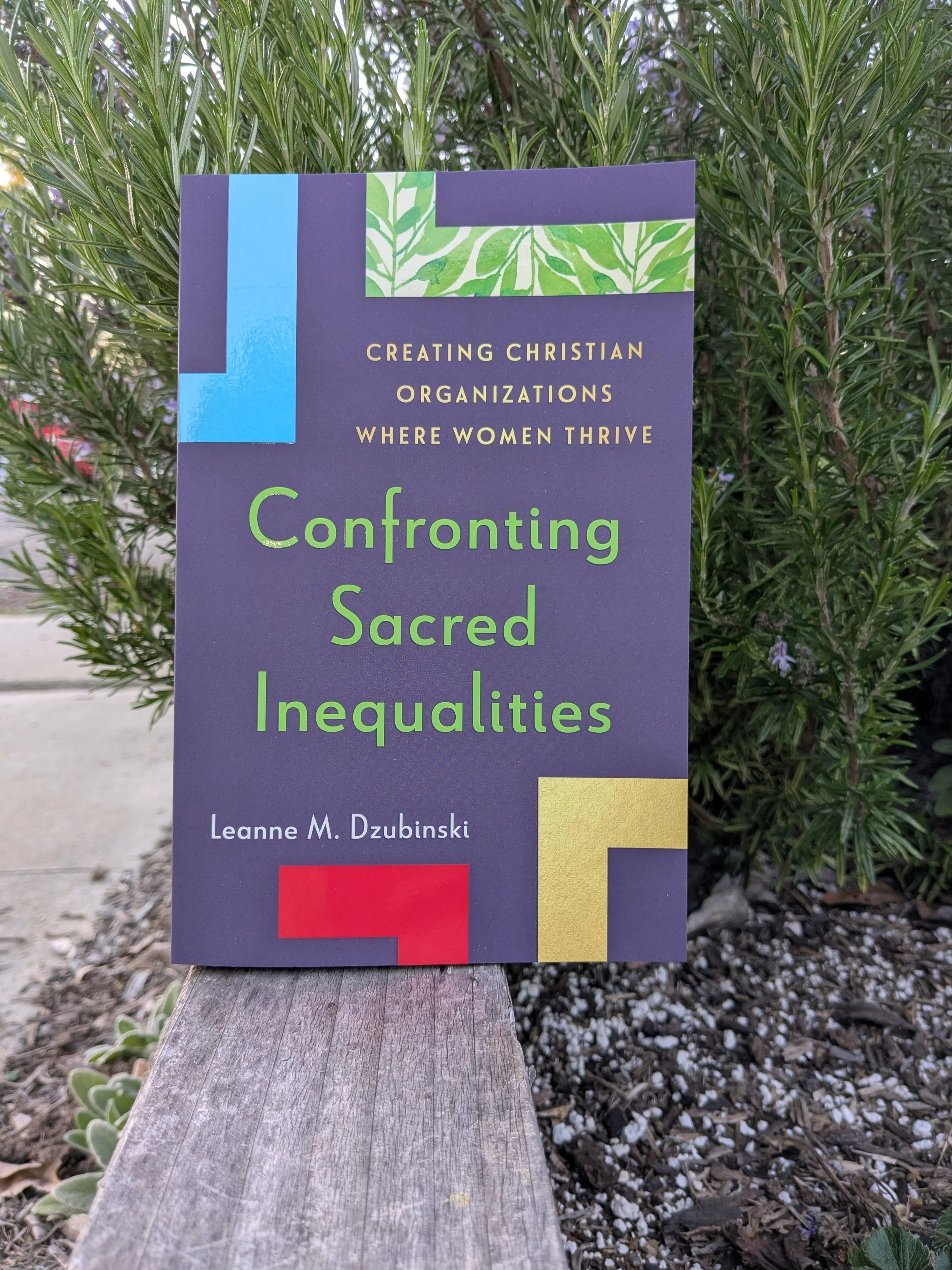 Book titled 'Confronting Sacred Inequalities' by Leanne M. Dzubinski, with a subtitle about creating Christian organizations where women thrive, placed outdoors on a wooden surface with green foliage in the background.