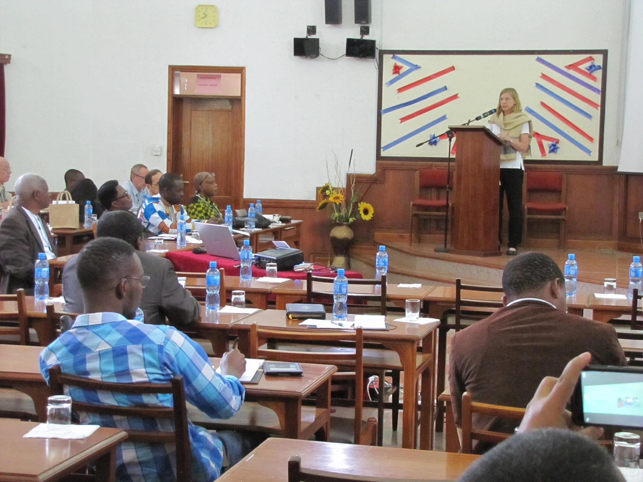 A woman giving a presentation at a conference or seminar, standing at a podium on a stage with a microphone, audience members seated at tables, some taking notes or pictures, bottles of water on the tables, and a decorated background behind the speaker.