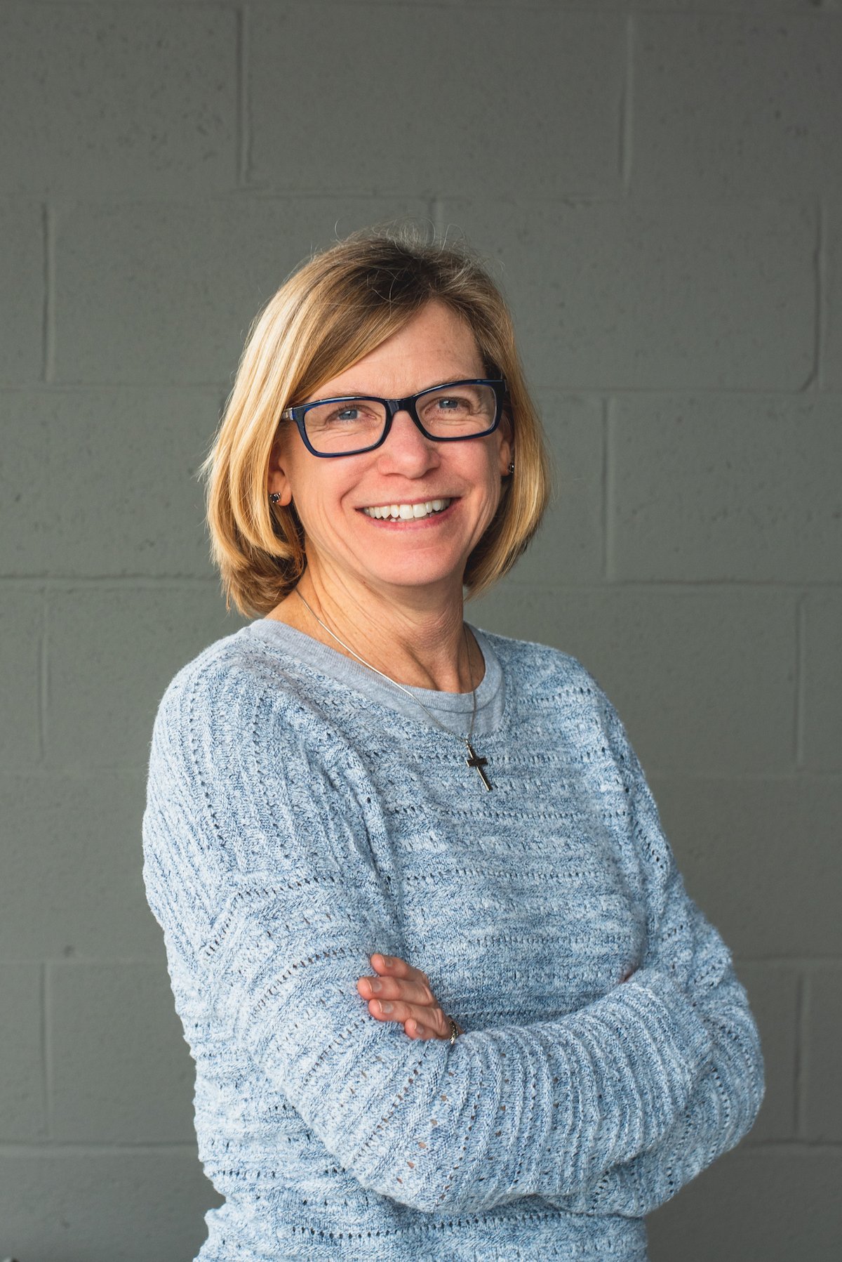A woman with short blonde hair, glasses, and a gray sweater standing against a plain gray wall, smiling with her arms crossed.