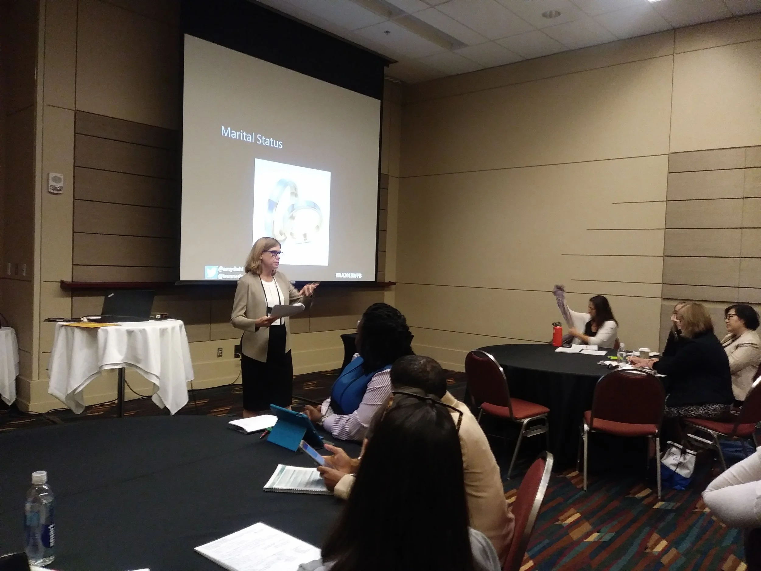 A woman giving a presentation in a conference room with seated attendees watching and taking notes