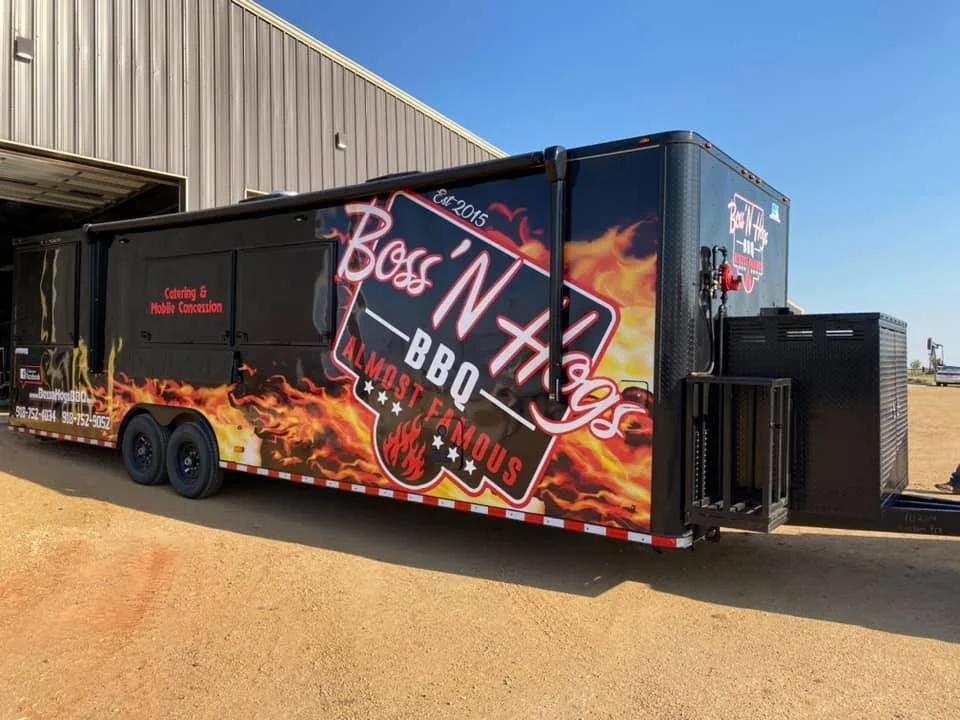 A black barbecue food truck with vibrant flame and fire graphics, featuring the business name "Boss 'N' Togs BBQ" and the tagline "Most Famous." The truck is parked outside near a building with an open garage, under a clear blue sky.