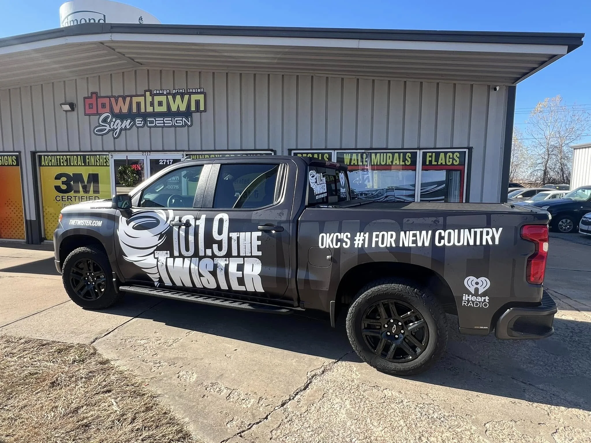 Black pickup truck with promotional vinyl wrap for radio station 101.9 The TWISTER parked in front of downtown sign. The truck features the station's logo, slogan, and website, along with logos for iHeartRadio and mentions of country music branding.