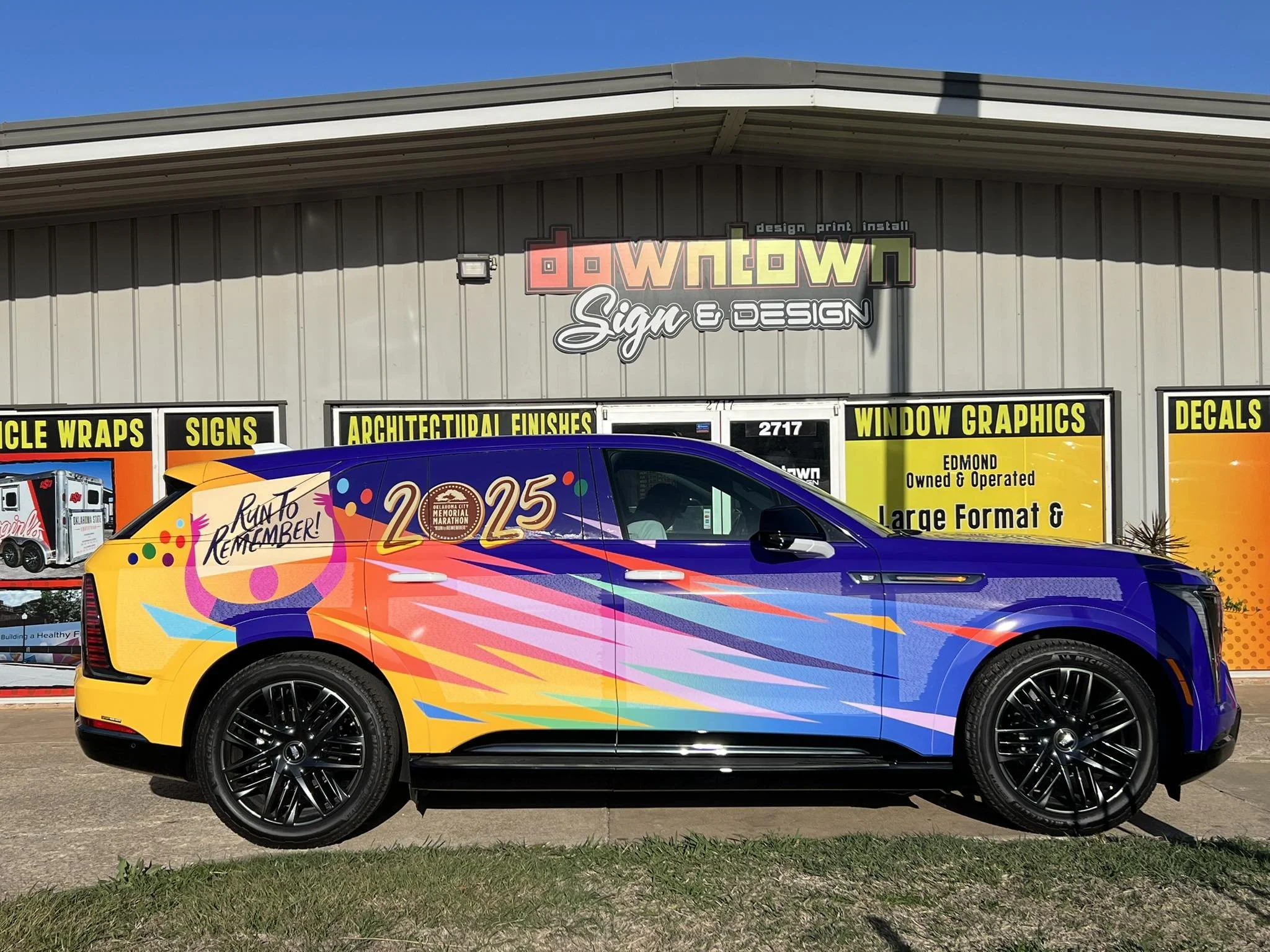 A colorful vehicle with 2025 and Run To REMEMBER! written on the side, parked in front of a sign for a sign and design shop.