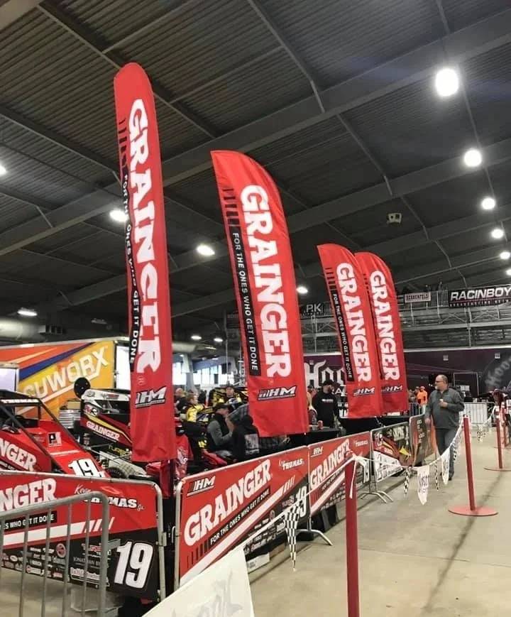 Indoor racing event with red feather flags displaying the name 'GRAINGER' and racing cars behind a barrier. Designed and printed by downtown sign and design