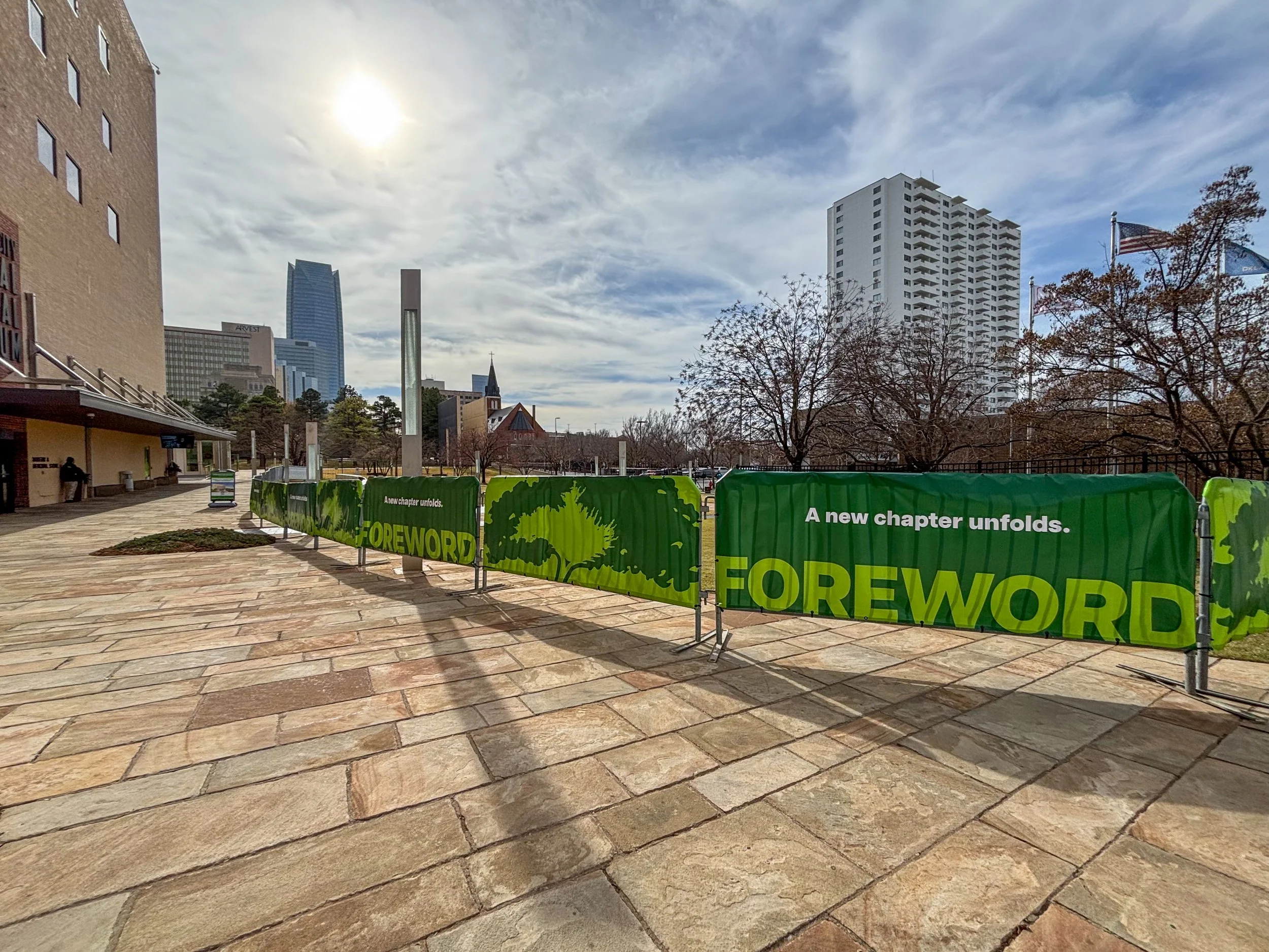 Large banner with vibrant colors reading 'Congrats Runners 2021 RUN to REMEMBER, Oklahoma City Memorial Marathon'. The banner features cartoon characters and various logos, with people in the background and city skyscrapers behind.