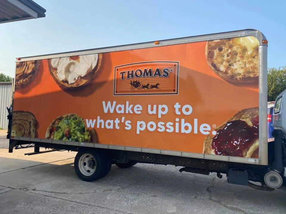 A food truck with a large orange advertisement displaying images of baked goods and the slogan "Wake up to what's possible". The truck is branded with Thomas' bakery logo.