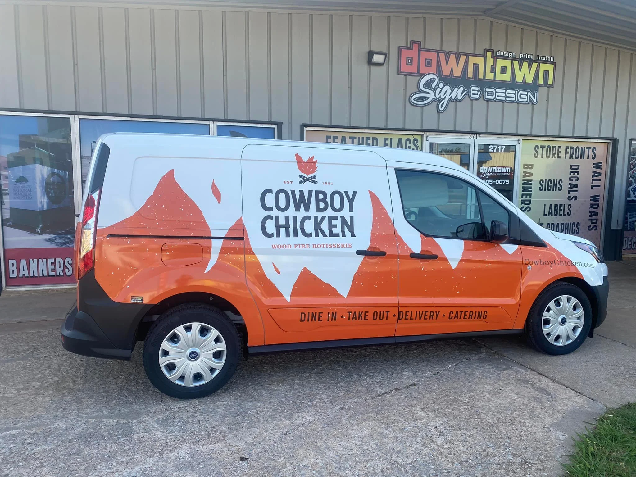 A delivery van parked outside a sign shop, displaying a graphic of a mountain with a chicken silhouette and the text 'Cowboy Chicken', advertising wood-fire rotisserie chicken with options for dine-in, takeout, delivery, and catering.