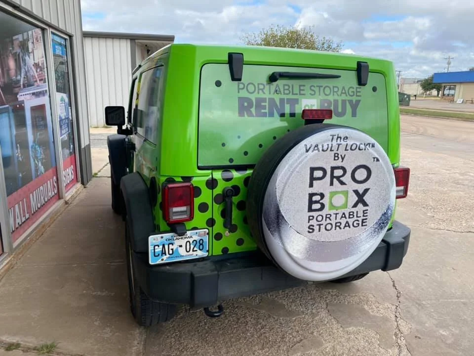 Green and black Pro Box portable storage vehicle with a spare tire cover that displays the brand logo, parked outside a building with signs for wall murals and signs in a small-town area.