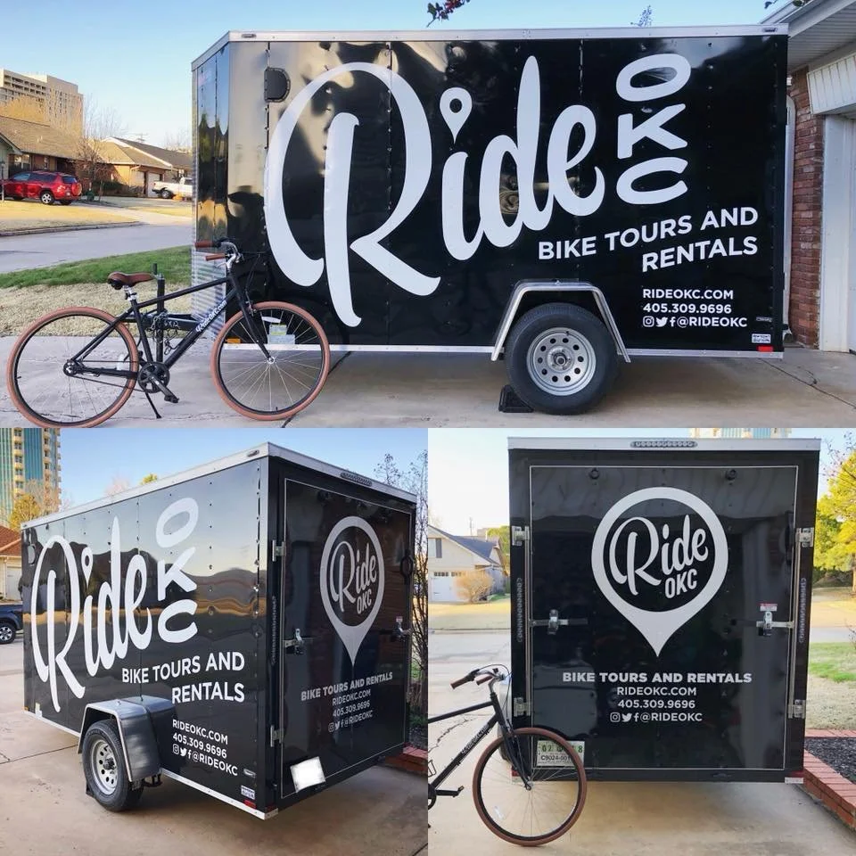 A black trailer with white lettering and logo for Ride OKC bike tours and rentals, parked outside a house. A black bicycle with brown tires is chained to the trailer. The trailer features the company's website, phone number, and social media handles.