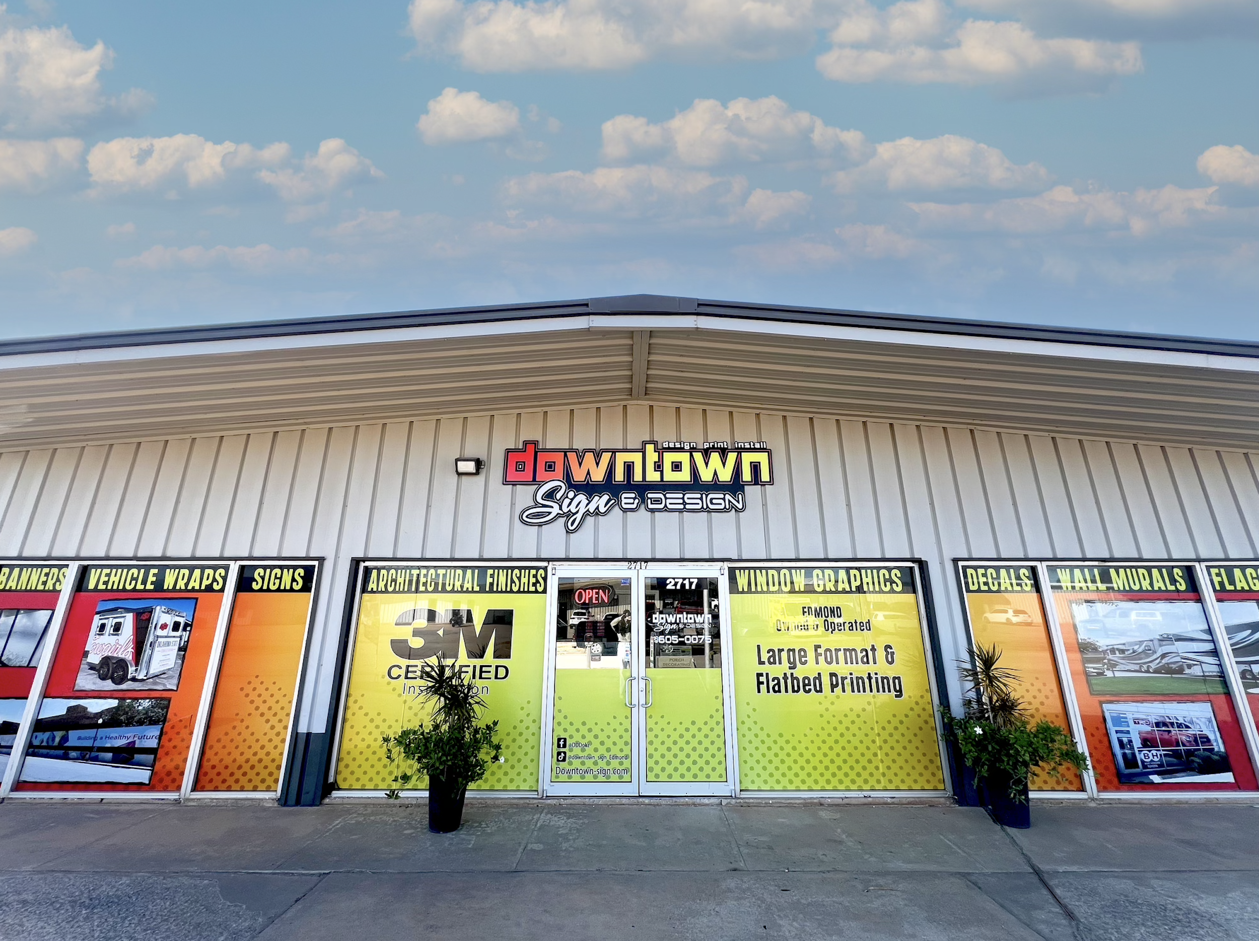 The storefront of a signage business called Downtown Signs & Design, featuring advertisements for vehicle wraps, signs, architectural finishes, window graphics, decals, wall murals, and flags, with an 'Open' sign on the door and plants outside.