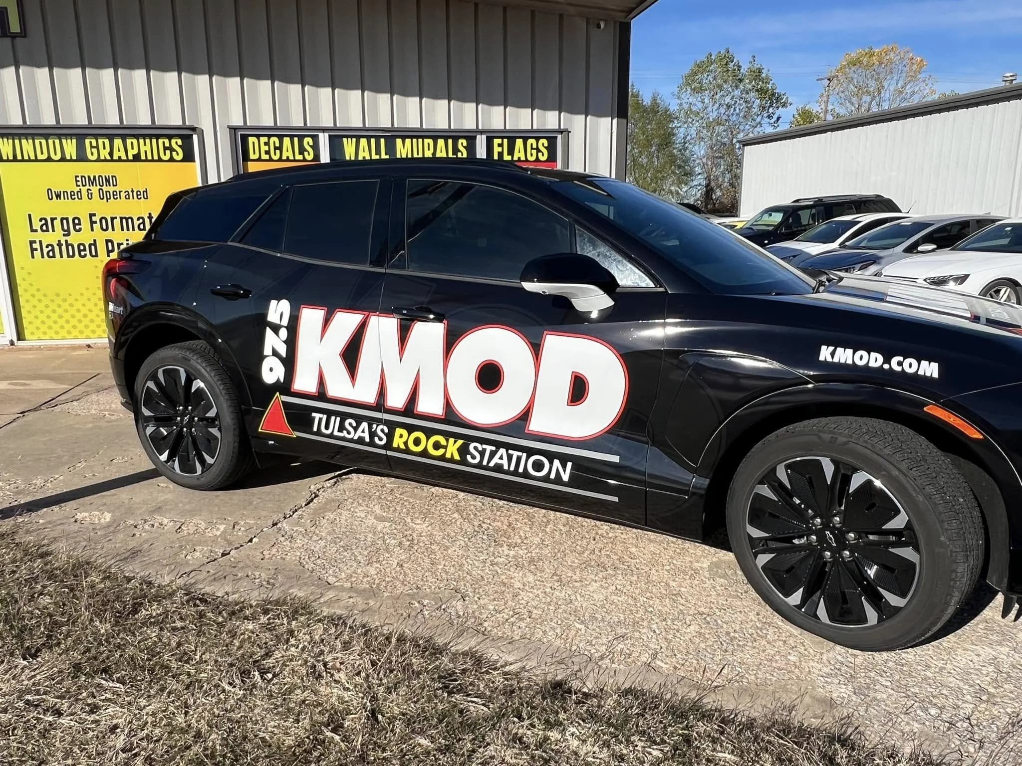 Black car with large promotional signage for 97.5 KMOD on the side, parked in front of a building with yellow and black sign advertising window graphics, decals, wall murals, and flags. Multiple white cars are in the background.