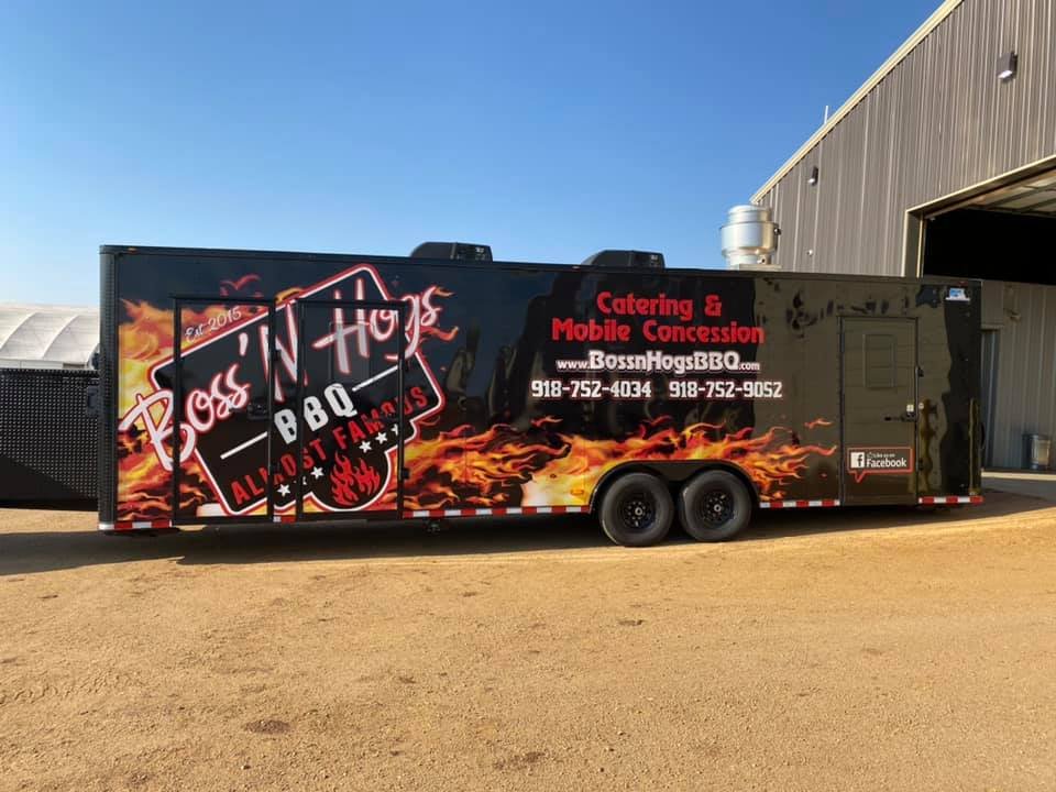 Black barbecue trailer with fiery graphics and the text 'Boss Hog BBQ' and 'ALMOST FAMOUS.' It offers catering and mobile concession services, with contact information and a Facebook logo.