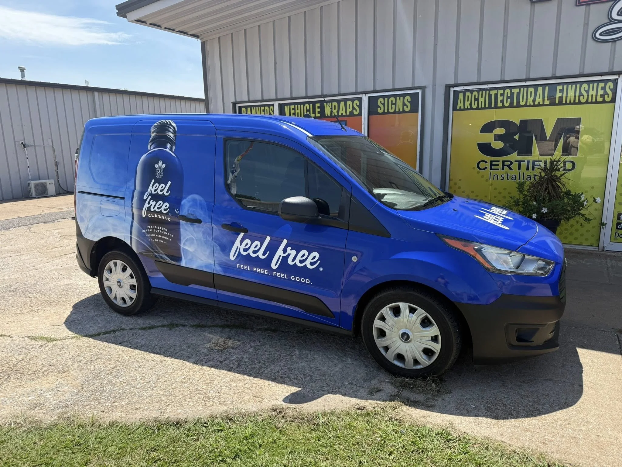 Blue commercial van with advertising for a herbal supplement called 'Feel Free' parked outside a building with signage for vehicle wraps, signs, and architectural finishes.
