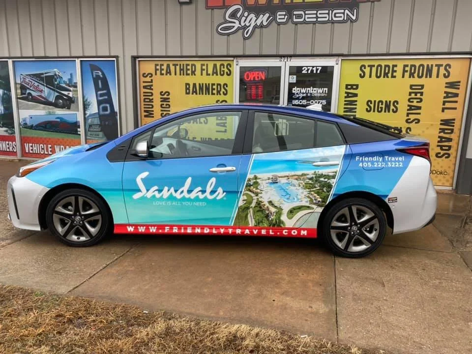 A car with a colorful wrap advertisement for Sandals travel company, parked in front of a store that sells signage and banners. The wrap features the Sandals logo, a scenic resort image, and the website www.friendlytravel.com.