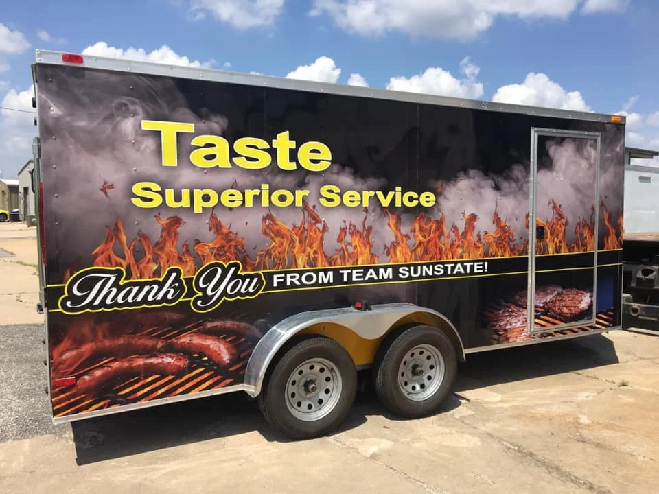 A food trailer with a graphic of flames and BBQ meats, including sausages and ribs, and text that says "Taste Superior Service. Thank You From Team Sunstate!"
