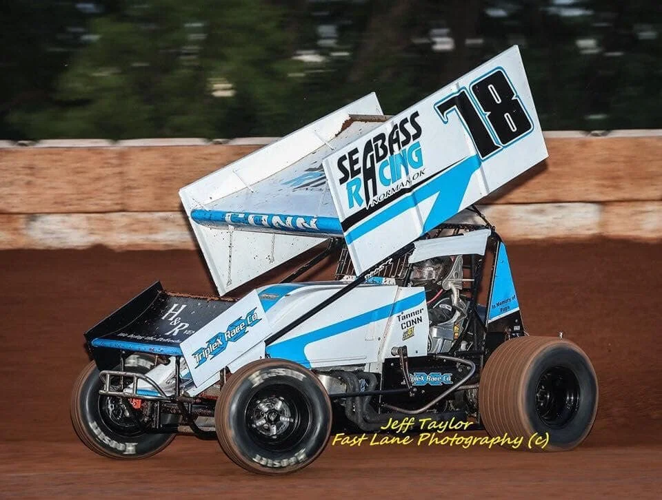 A blue and white sprint car racing on a dirt track, with a large wing on top and the number 18 on the side.