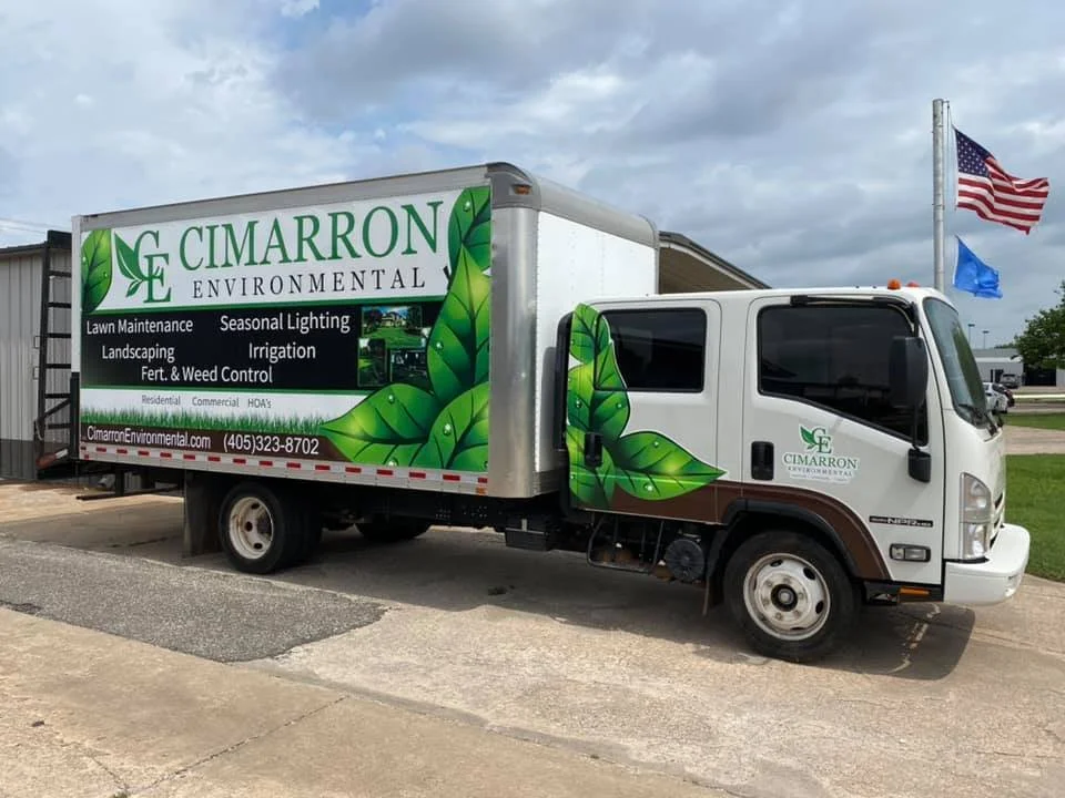 A white company truck with green leaf graphics parked on a street near a building, with American and blue flags flying in the background, advertising Cimarron Environmental services.