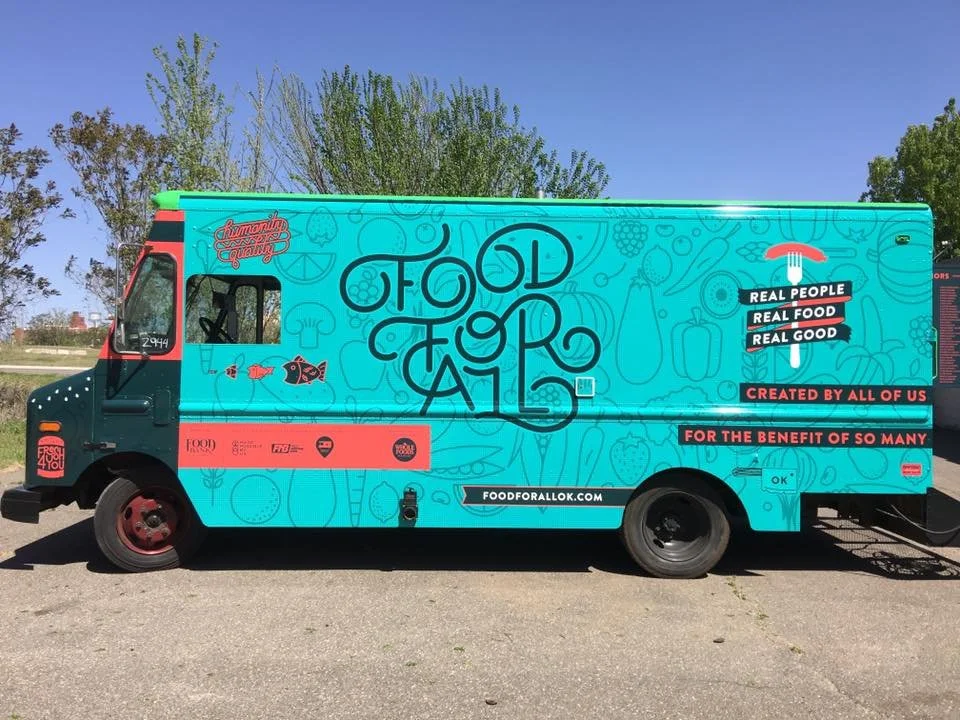 A colorful food truck with the words 'Food for All' and the slogan 'Real People, Real Food, Real Good.' The truck features illustrations of food items, fish, and kitchen utensils, and promotes community sharing and benefit.
