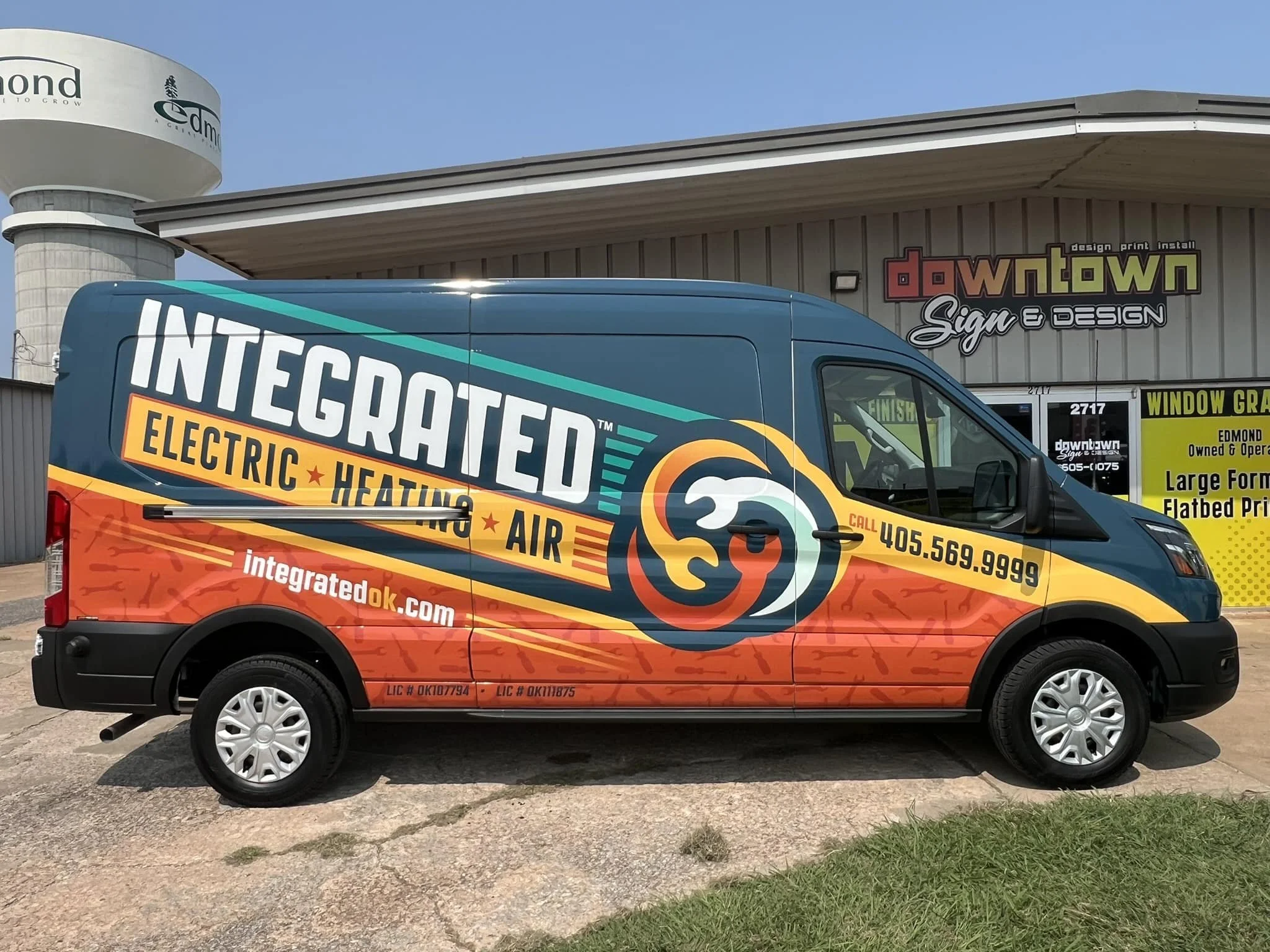 Company van for Integrated Electric Heating Air with a colorful design, contact phone number, website, and licensing details parked outside a signage and window graphics business.