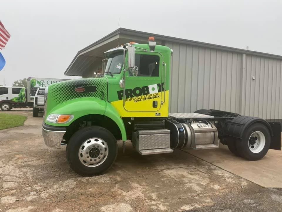 Green and yellow portable storage truck parked in front of a gray building with a partial American flag visible on the left.