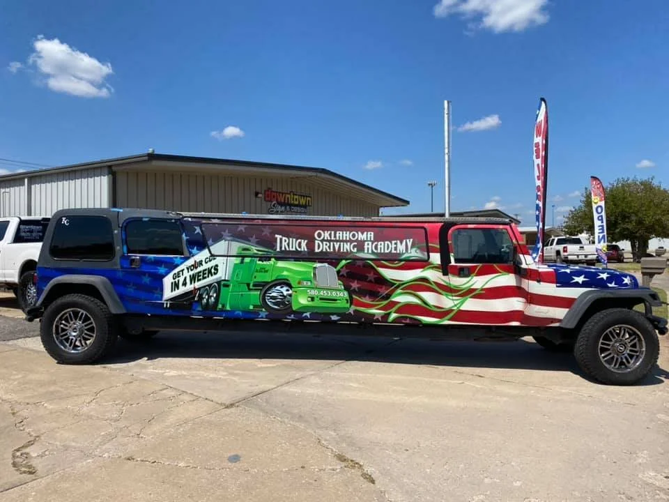 A long vehicle painted with an American flag and green trucks advertising the Oklahoma Truck Driving Academy. The vehicle has banners and flags nearby, with a building and parking lot in the background.